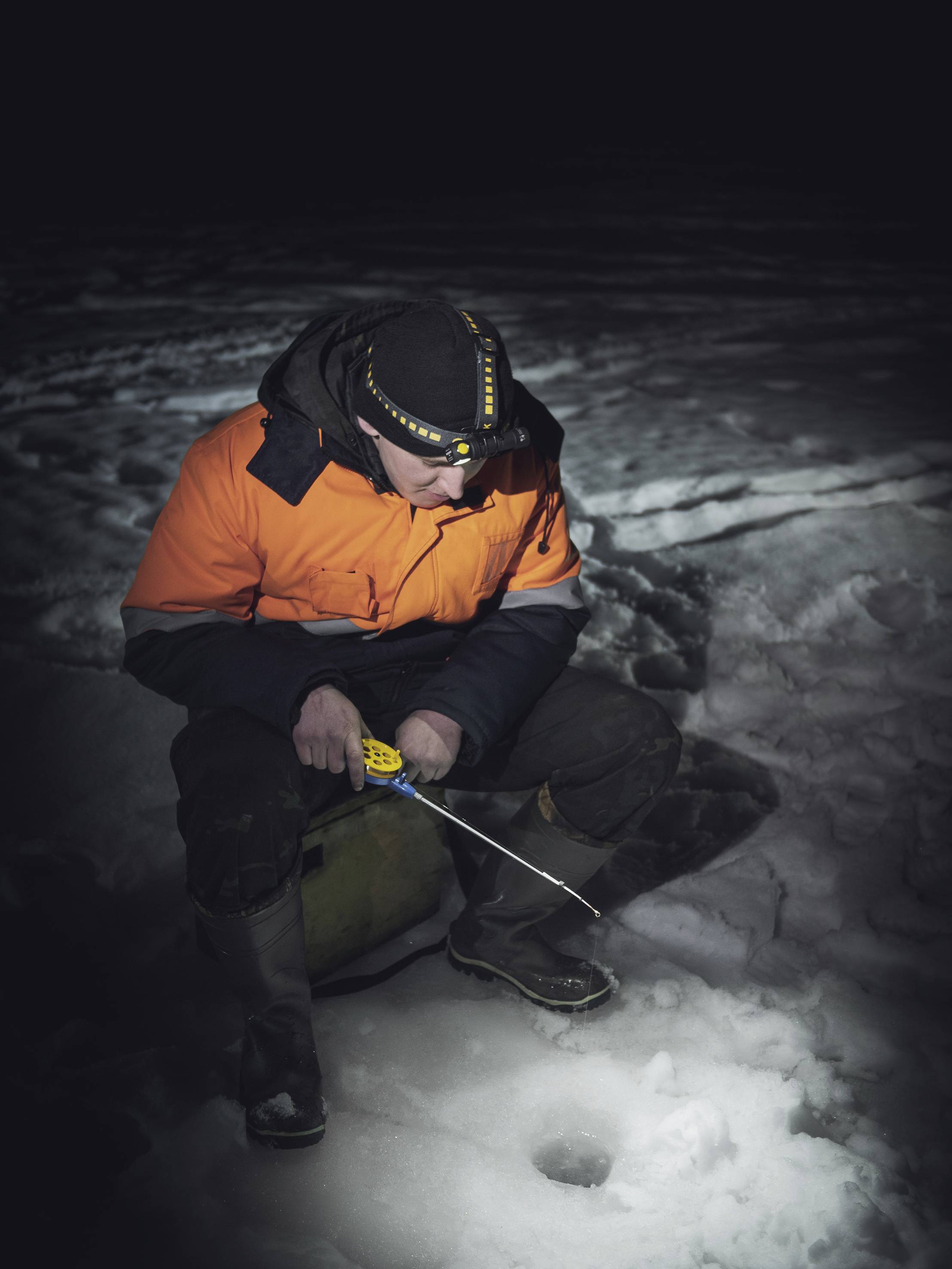 Eine Person in orangefarbener Jacke beim Eisfischen in der Nacht. Sie hält eine Angelrute und sitzt neben einem Eisloch im Schnee.
