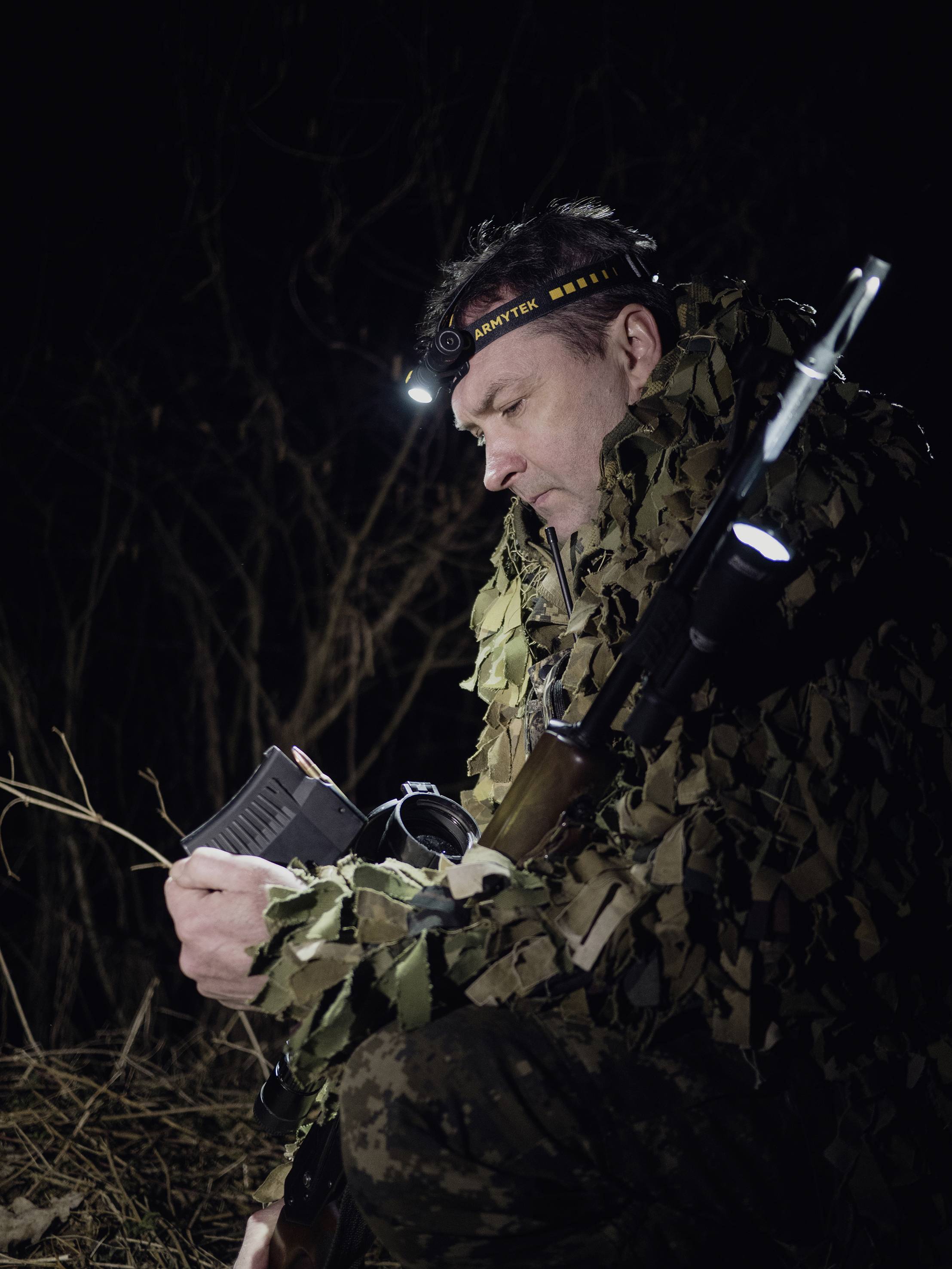 Eine Person in Tarnkleidung sitzt im Dunkeln im Freien und überprüft ein Magazin mit einer Stirnlampe. Sie hält ein Gewehr.