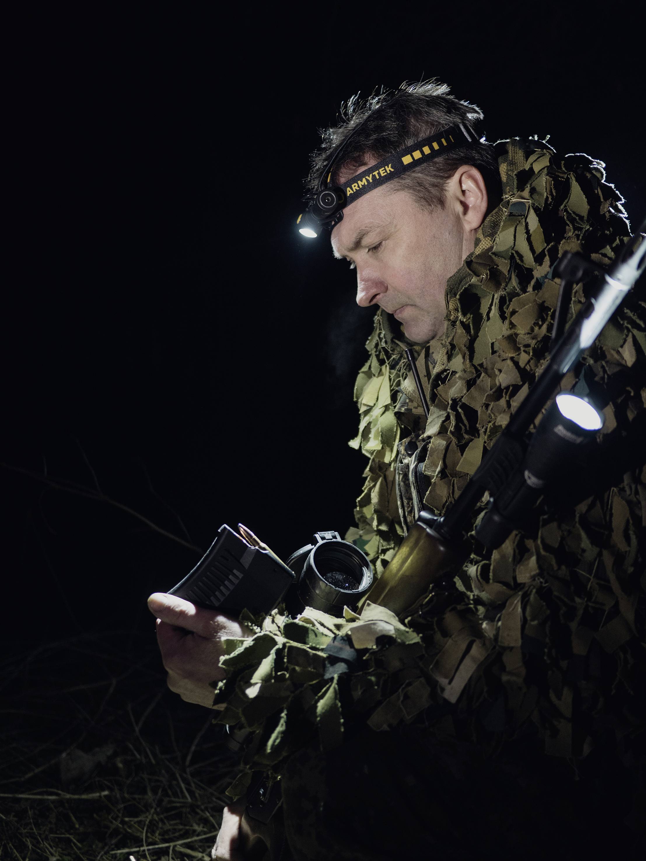 Eine Person in Tarnkleidung liest bei Nacht mit einer Stirnlampe in einem Notizbuch, während sie ein Gewehr trägt.