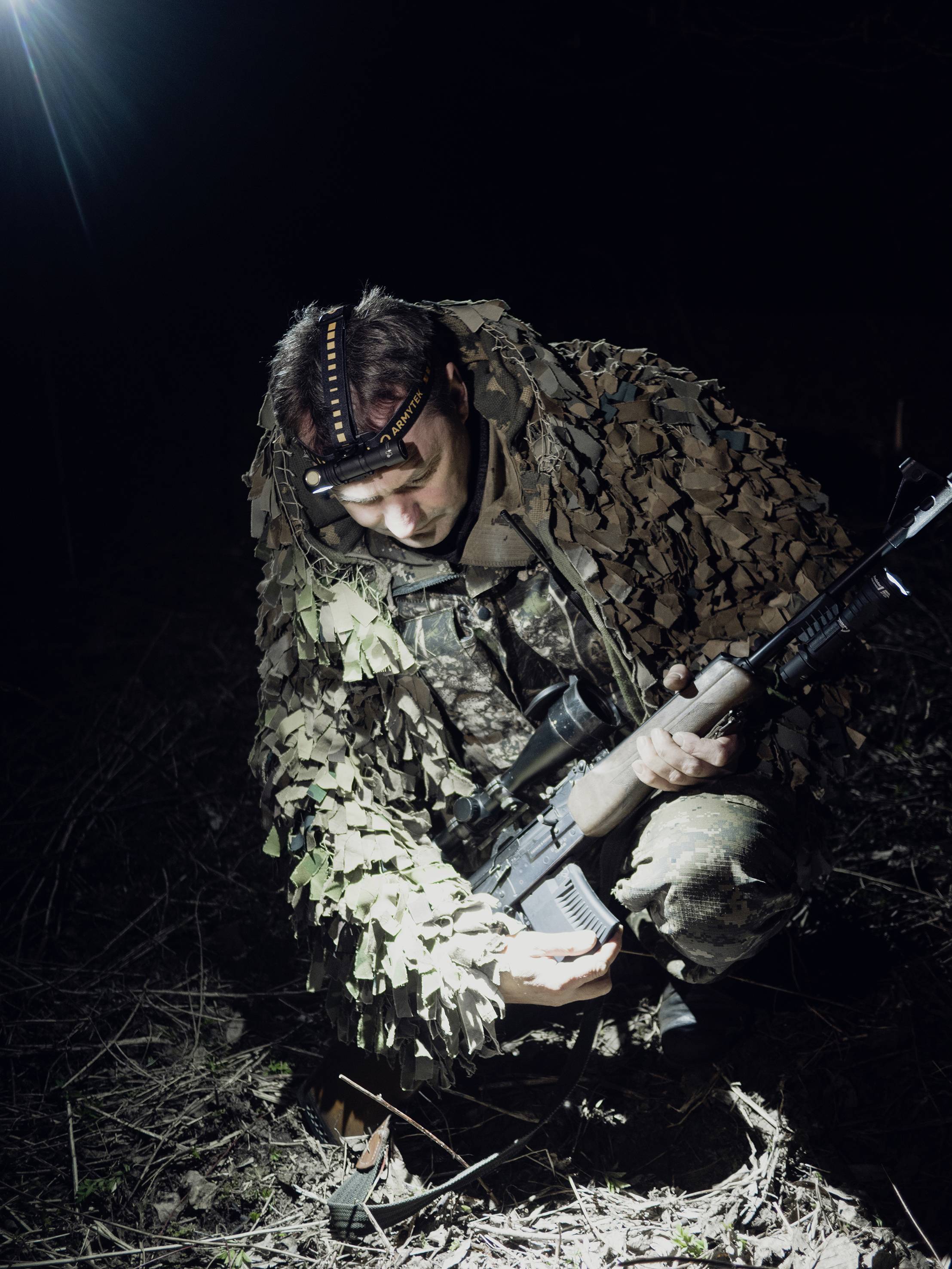Ein Soldat in Tarnkleidung kniet in einem dunklen Gelände und hält ein Gewehr, beleuchtet von einem starken Lichtstrahl.