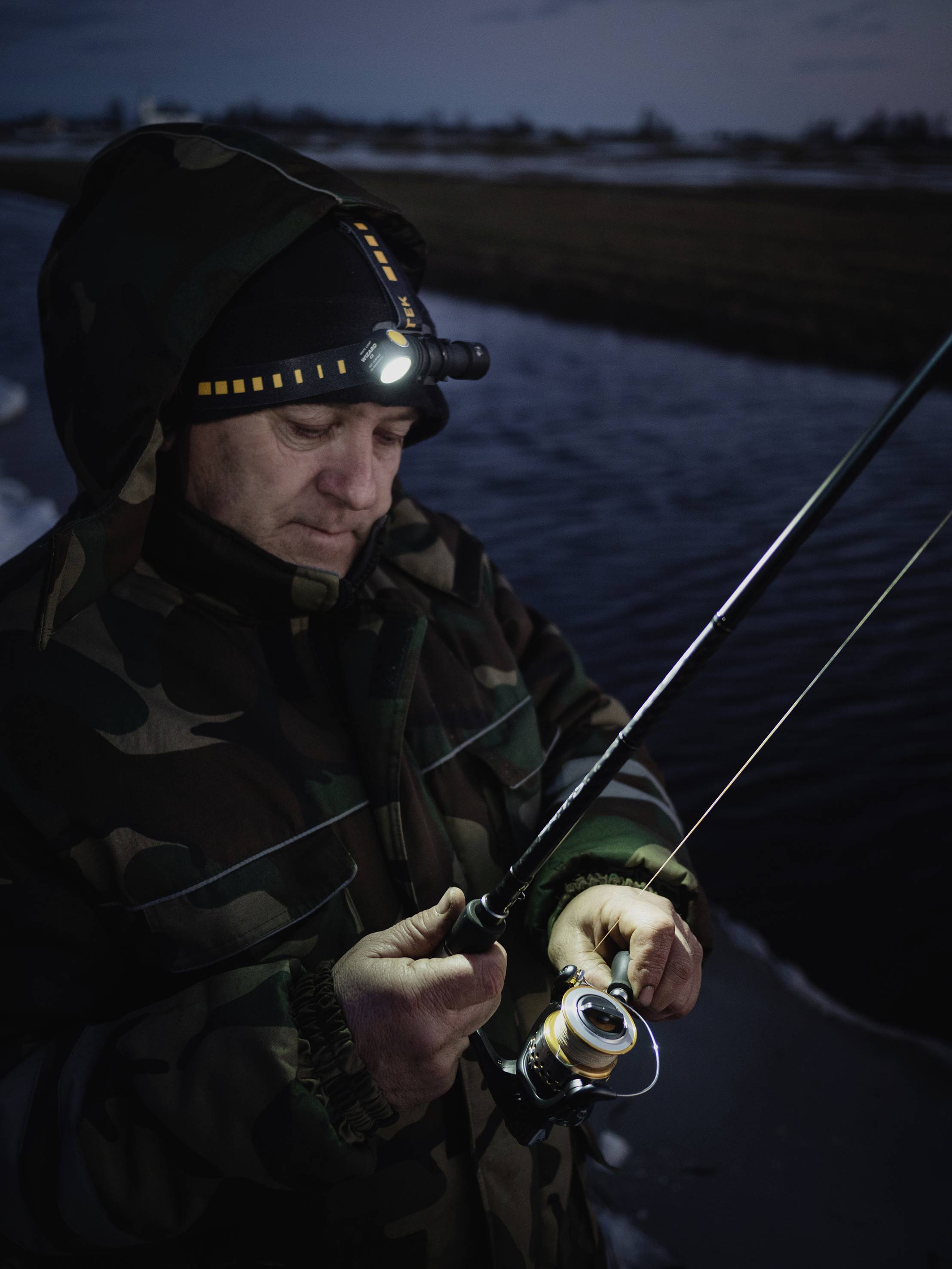 Ein Mann angelt bei Dämmerung am Fluss. Er trägt eine Tarnjacke und Stirnlampe. Trübe Beleuchtung, ruhige Atmosphäre.