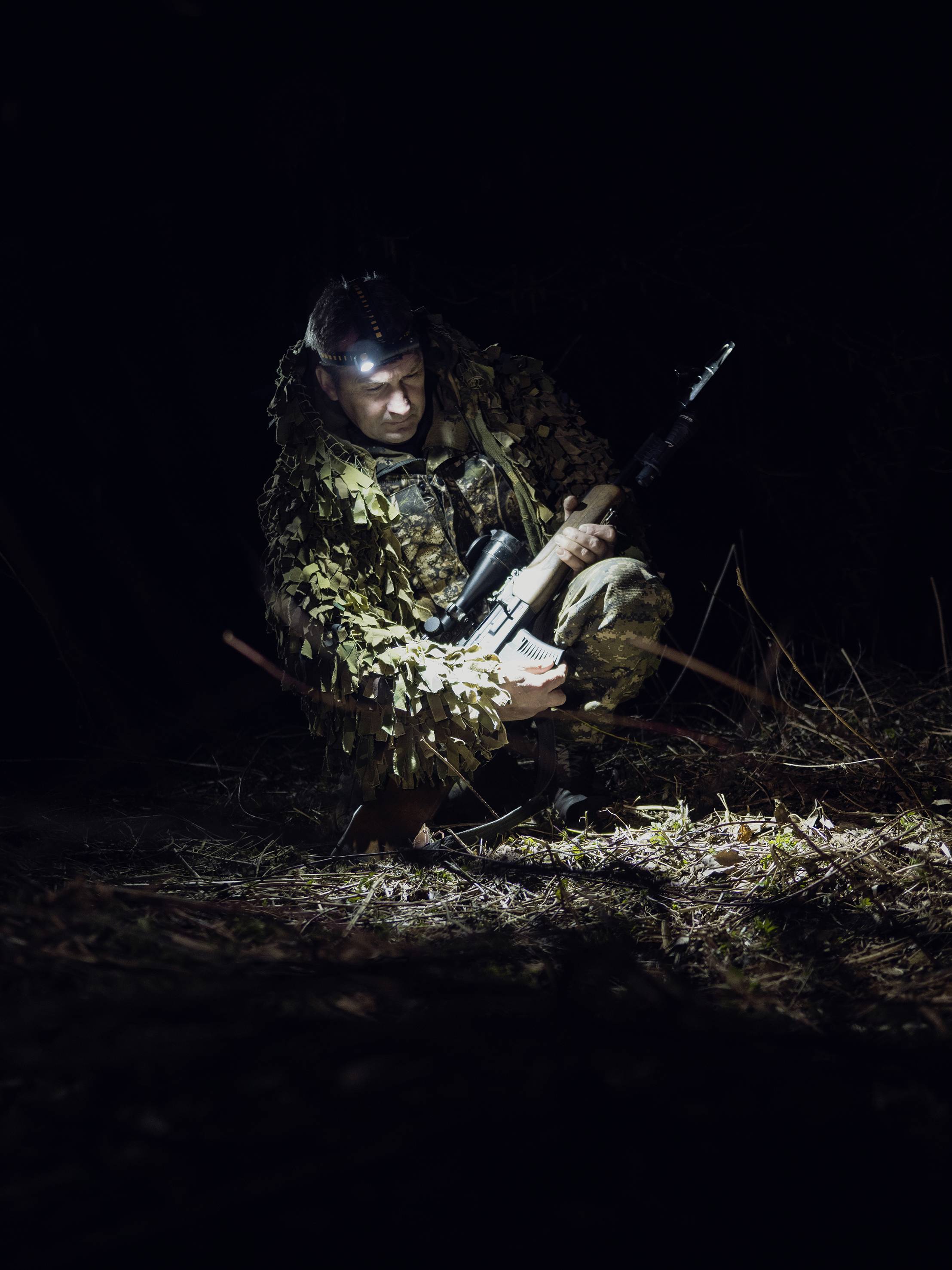 Ein Mensch in Tarnkleidung kniet nachts im Wald und hält ein Gewehr. Ein Scheinwerfer beleuchtet die Szene, während er den Boden betrachtet.