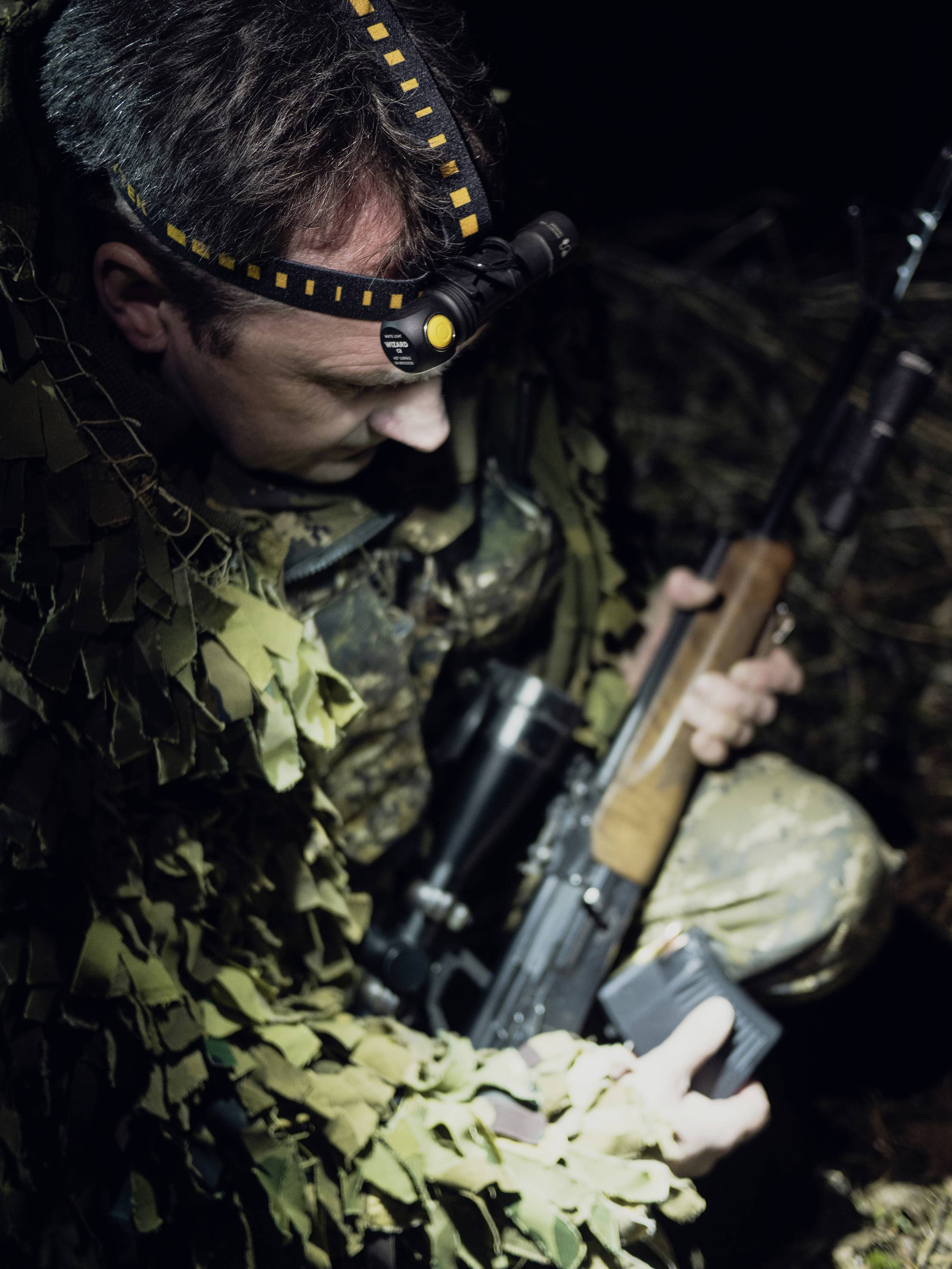 Eine Person in Tarnkleidung hält ein Gewehr und trägt eine Stirnlampe. Sie befindet sich offenbar in einem dämmerigen Waldgebiet.