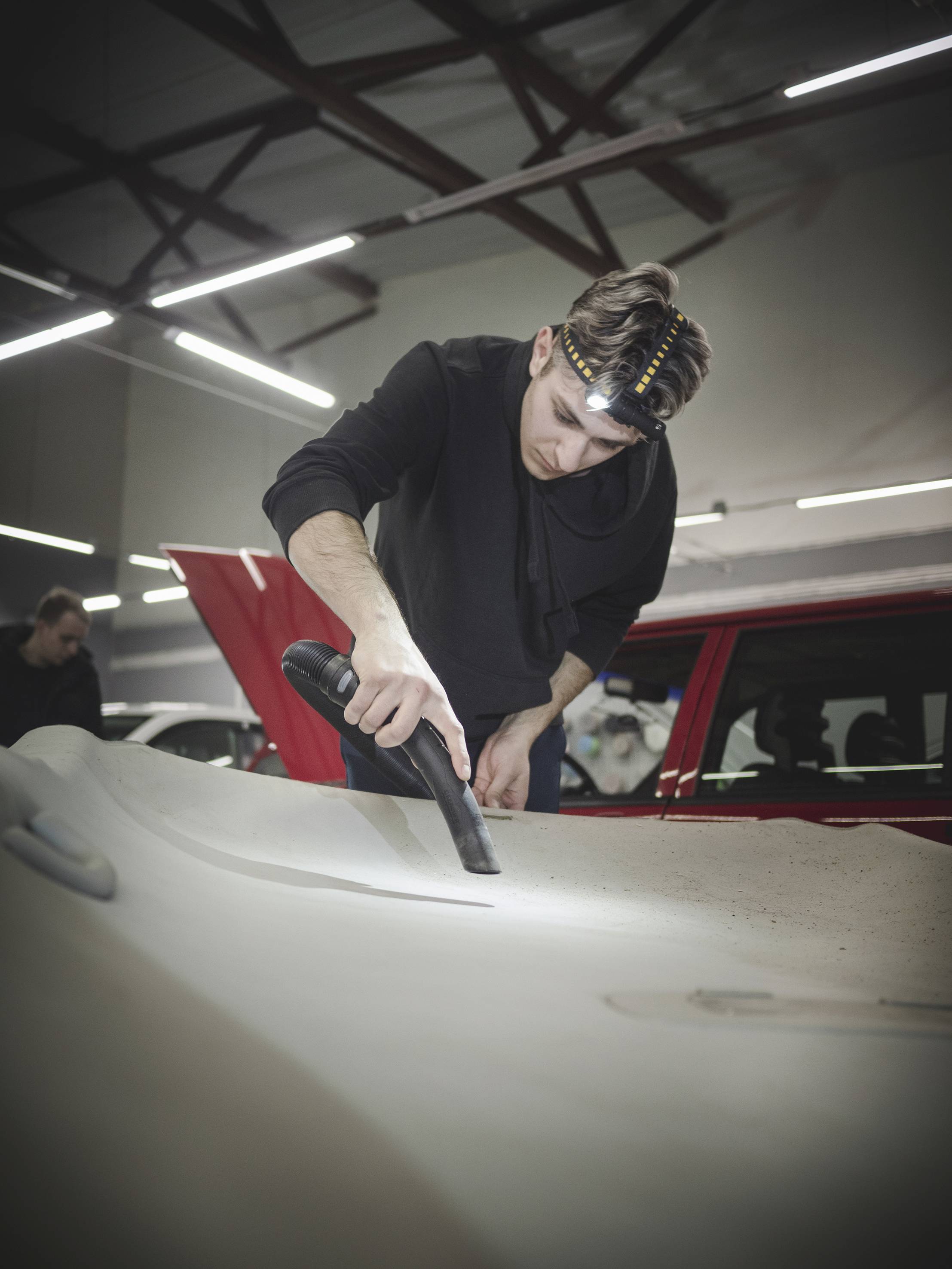 Ein Mann mit Stirnband saugt die Motorhaube eines Autos in einer Werkstatt ab. Im Hintergrund sind weitere Personen und ein rotes Auto zu sehen.