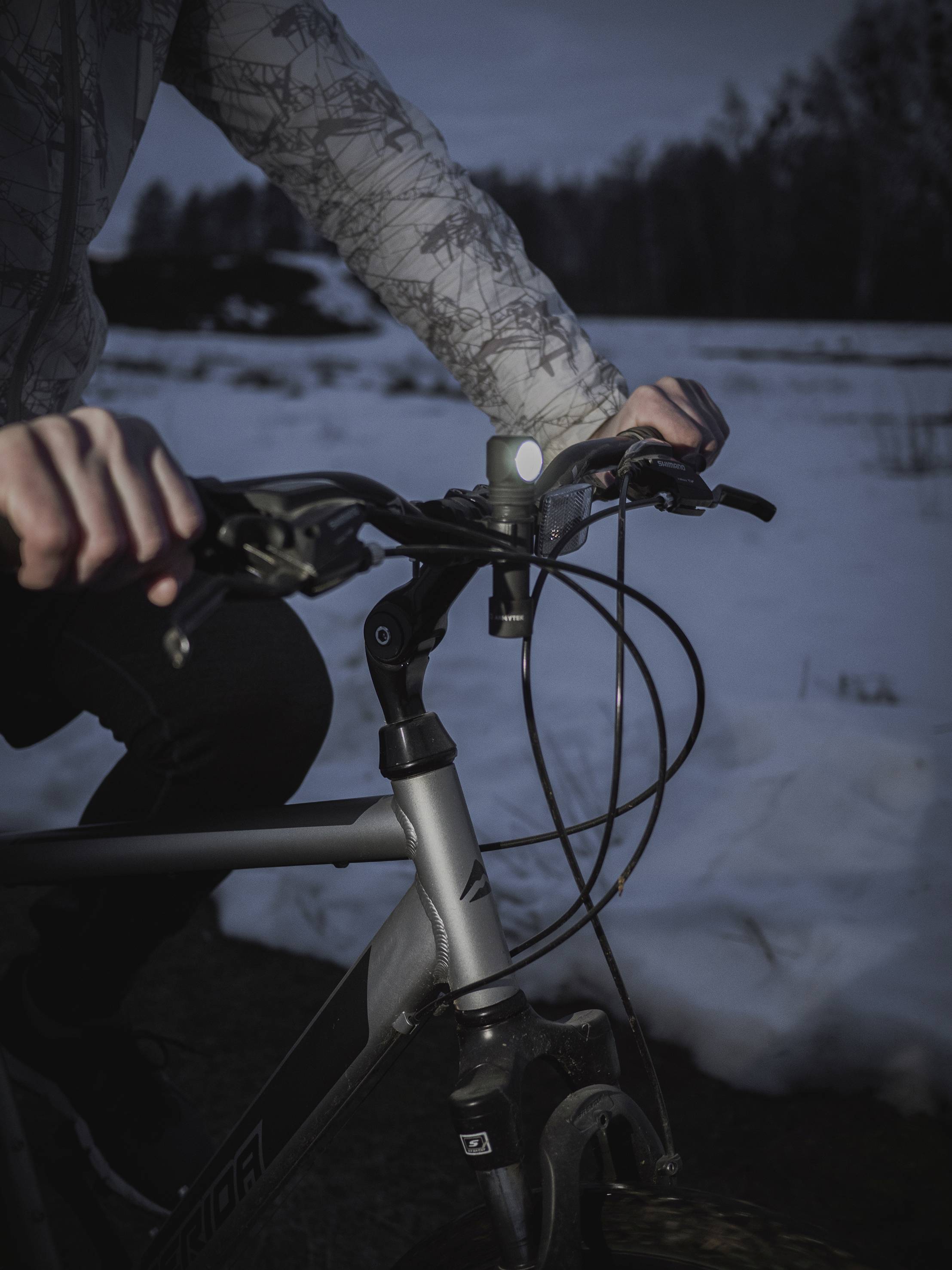 Person fährt bei Dämmerung ein Fahrrad auf verschneitem Weg. Fokus auf Lenker und Hände, Hintergrund unscharf mit Schnee.