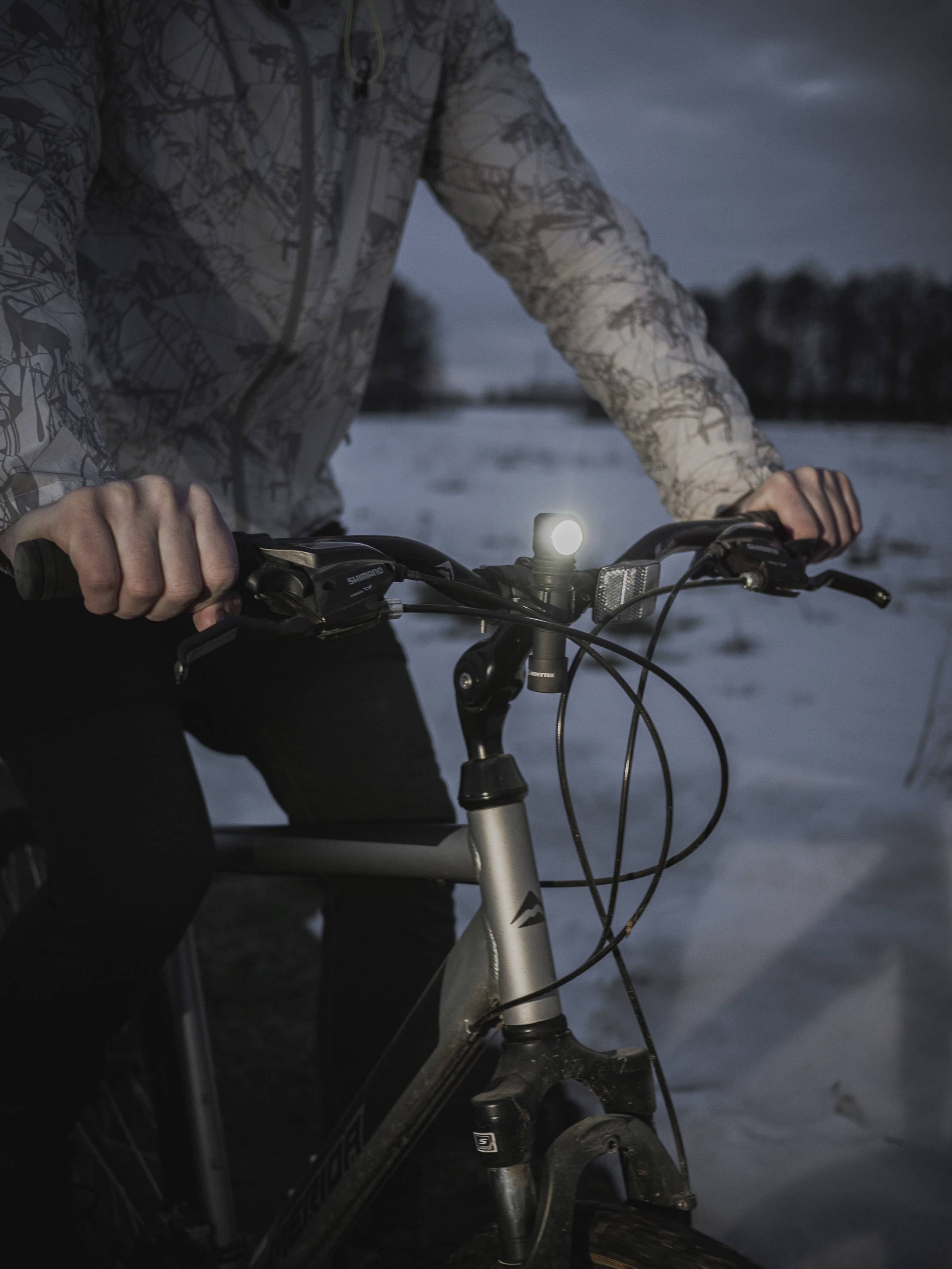 Eine Person fährt bei Dämmerung auf einem Fahrrad mit eingeschaltetem Licht durch eine schneebedeckte Landschaft.