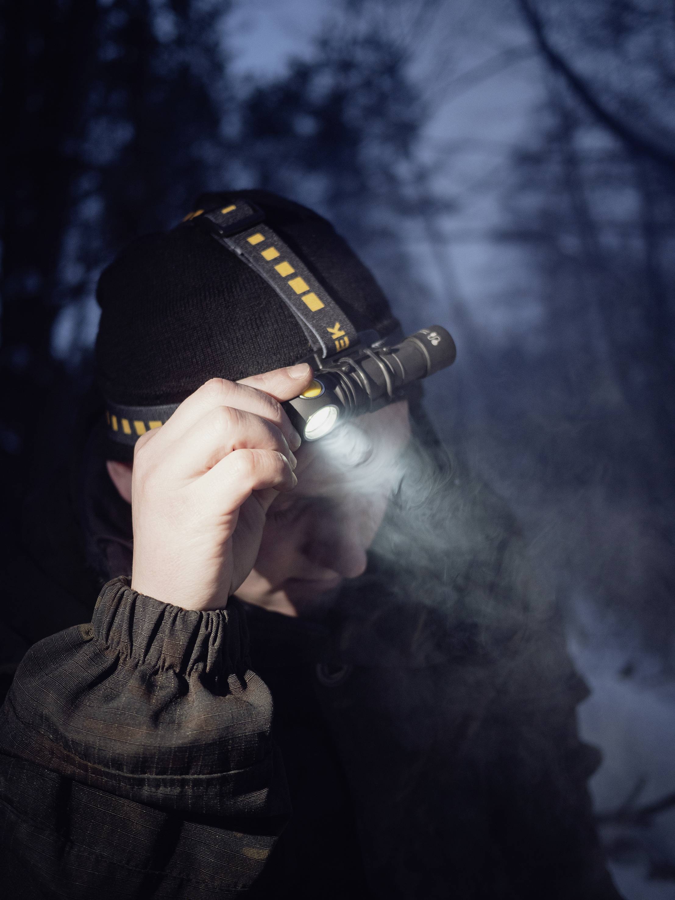Eine Person im Wald, gekleidet in dunkler Kleidung, hält eine leuchtende Stirnlampe an der Stirn, in die winterliche Dämmerung blickend.
