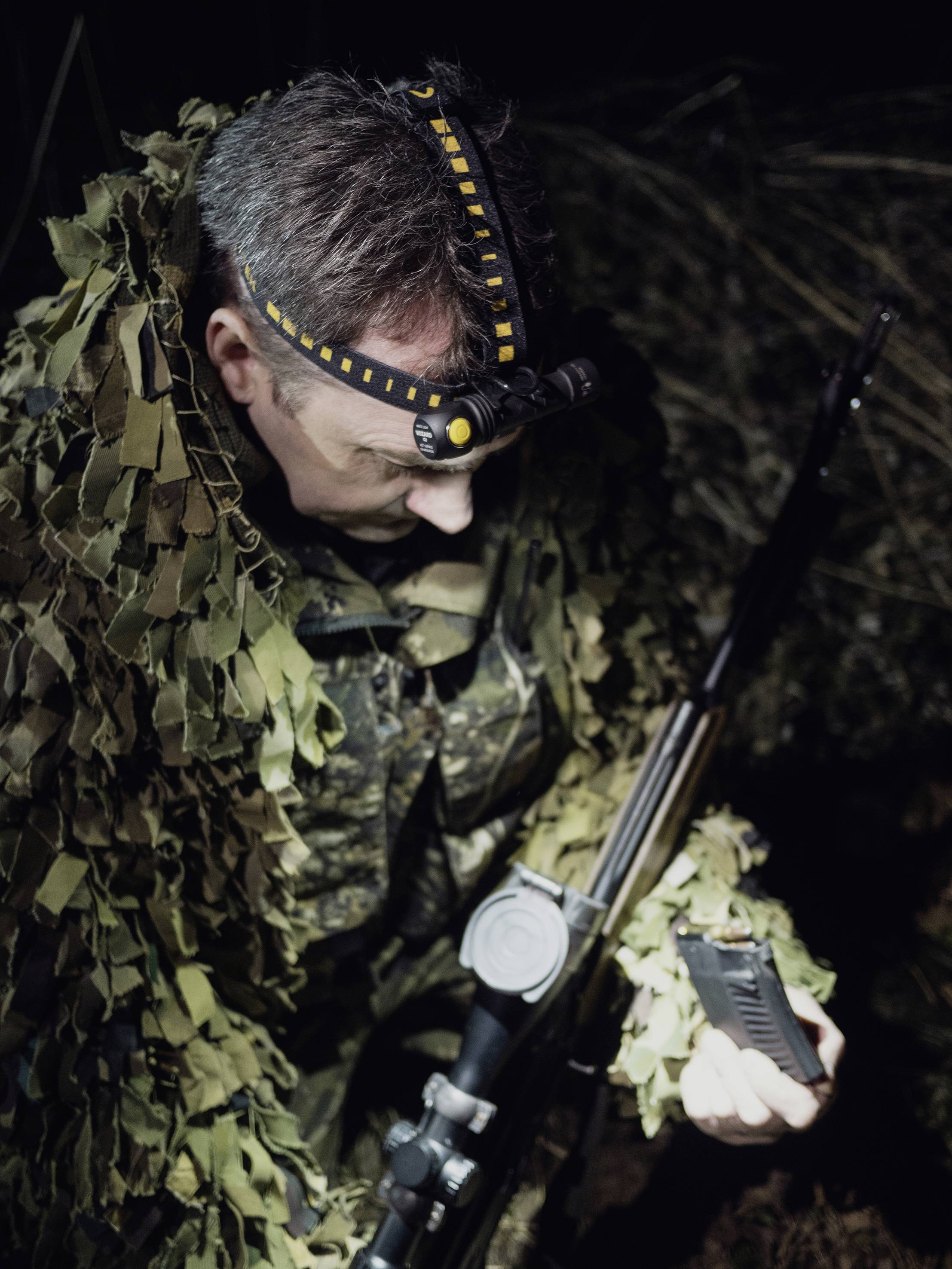 Eine Person in Tarnkleidung hält ein Gewehr und eine Munitionseinheit, vermutlich bei einer nächtlichen Jagd oder militärischen Übung.