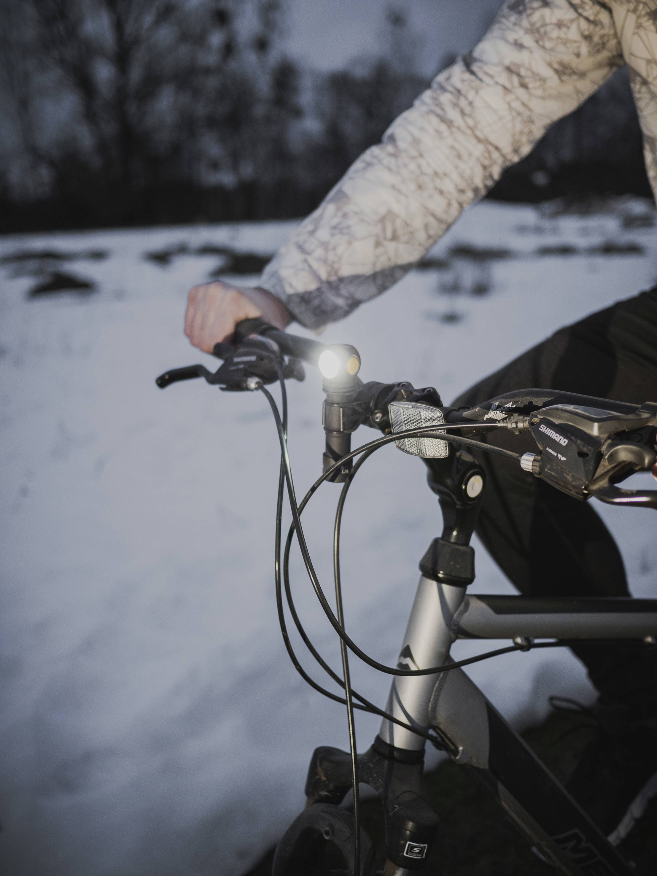 Person fährt ein Fahrrad im Schnee, trägt eine gemusterte Jacke. Das Fahrradlicht leuchtet, es ist abends oder früh morgens.