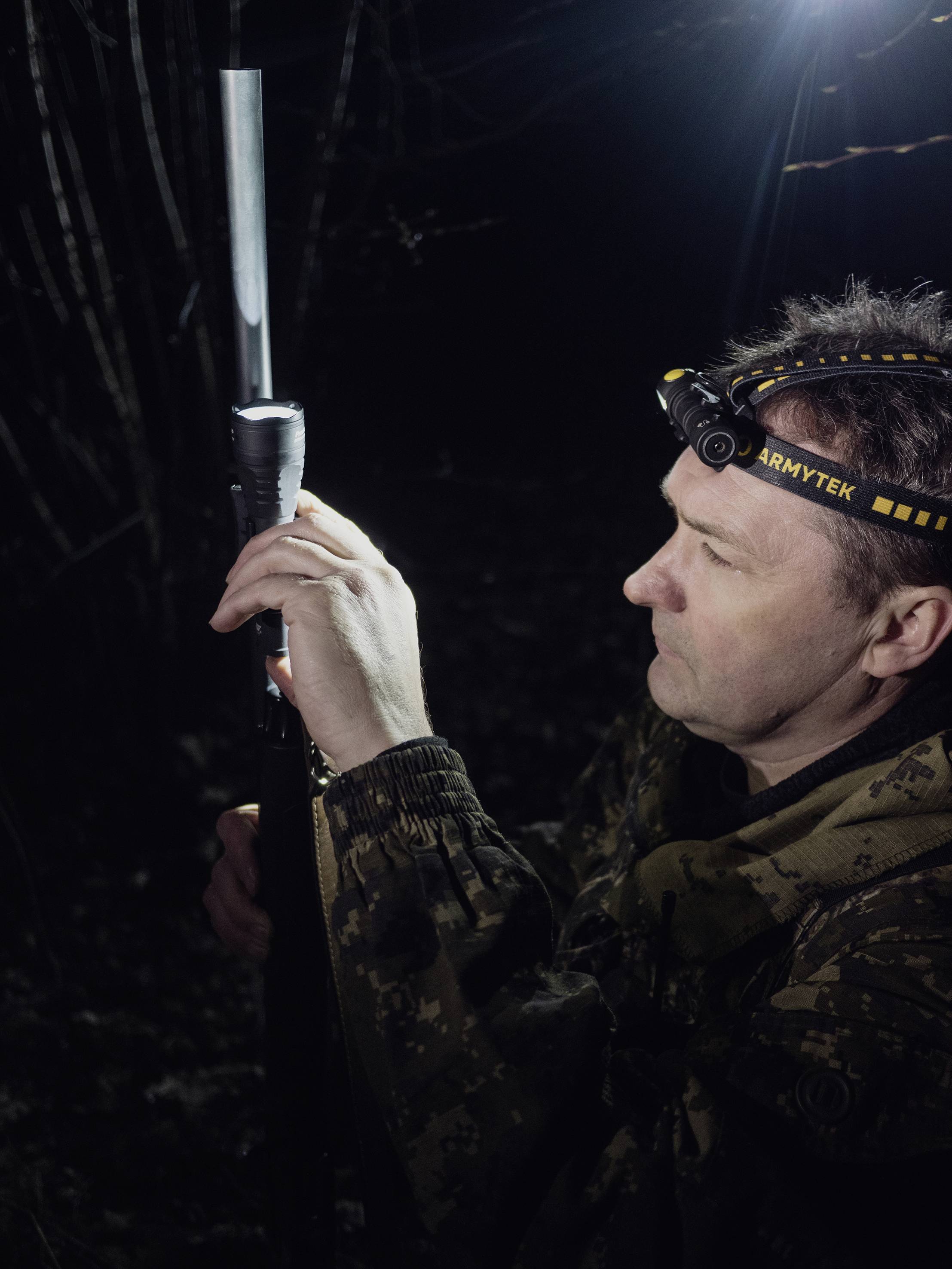 Mann im militärisch aussehenden Anzug im Dunkeln. Er hält eine längliche Ausrüstung und trägt ein Stirnband mit Licht.