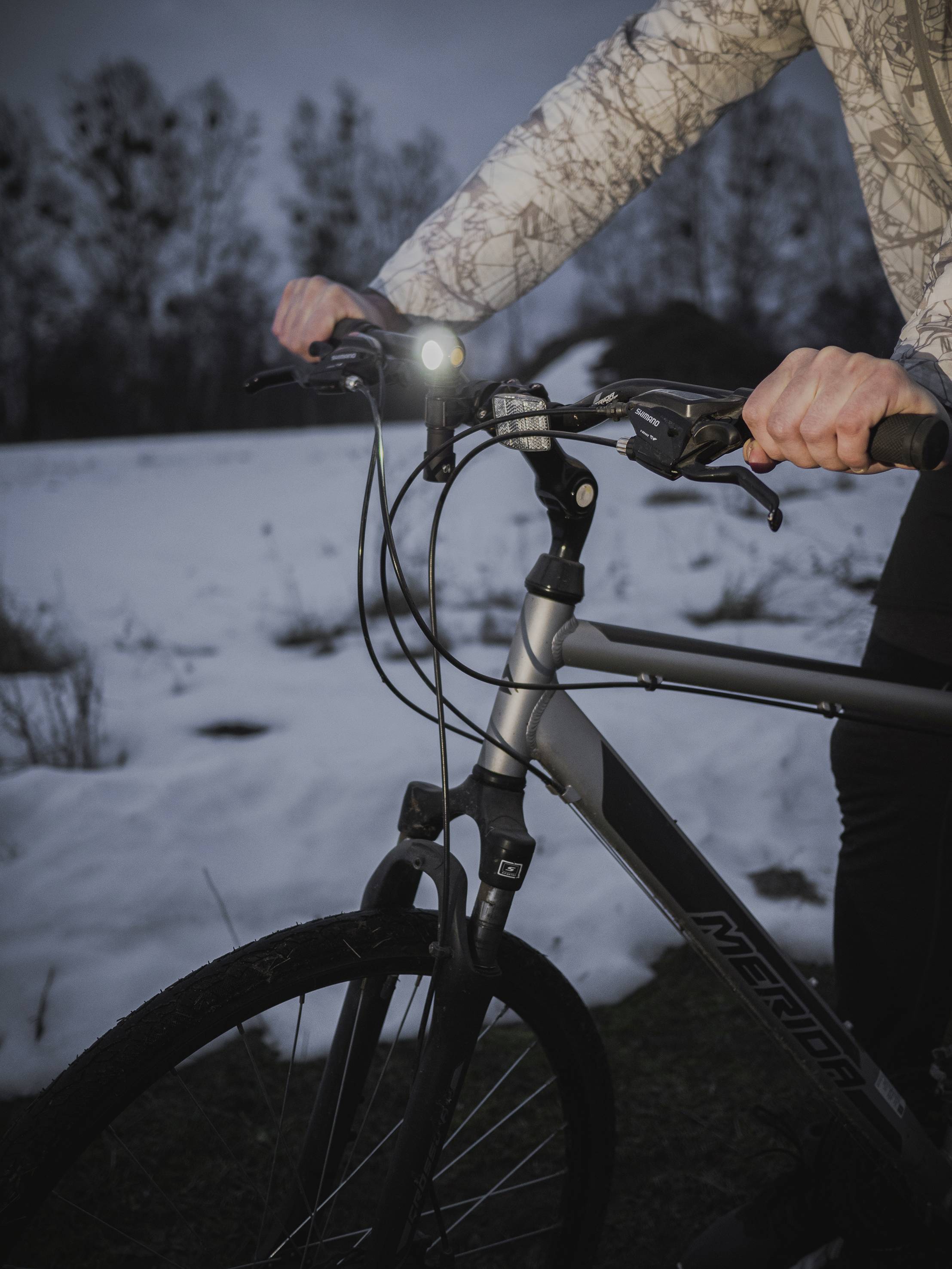 Eine Person hält den Lenker eines Mountainbikes im Freien. Der Schnee liegt auf dem Boden, es ist dämmrig. Fahrradlicht leuchtet.