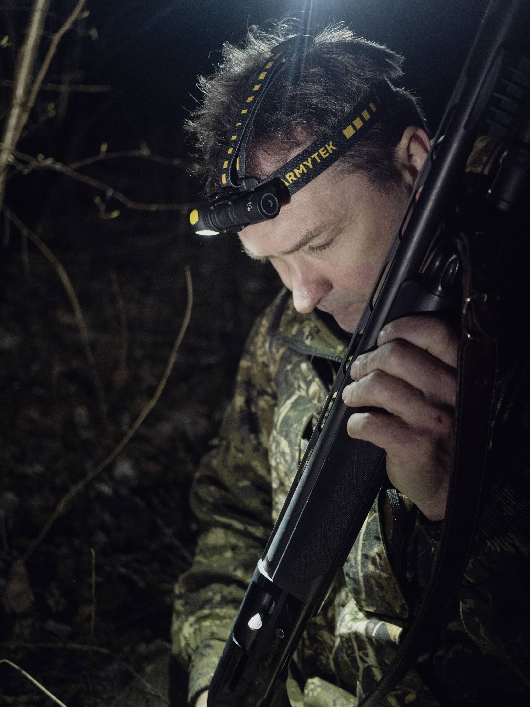 Ein Mann in Tarnkleidung hält ein Gewehr und trägt eine Stirnlampe, während er in einem dunklen Wald steht.