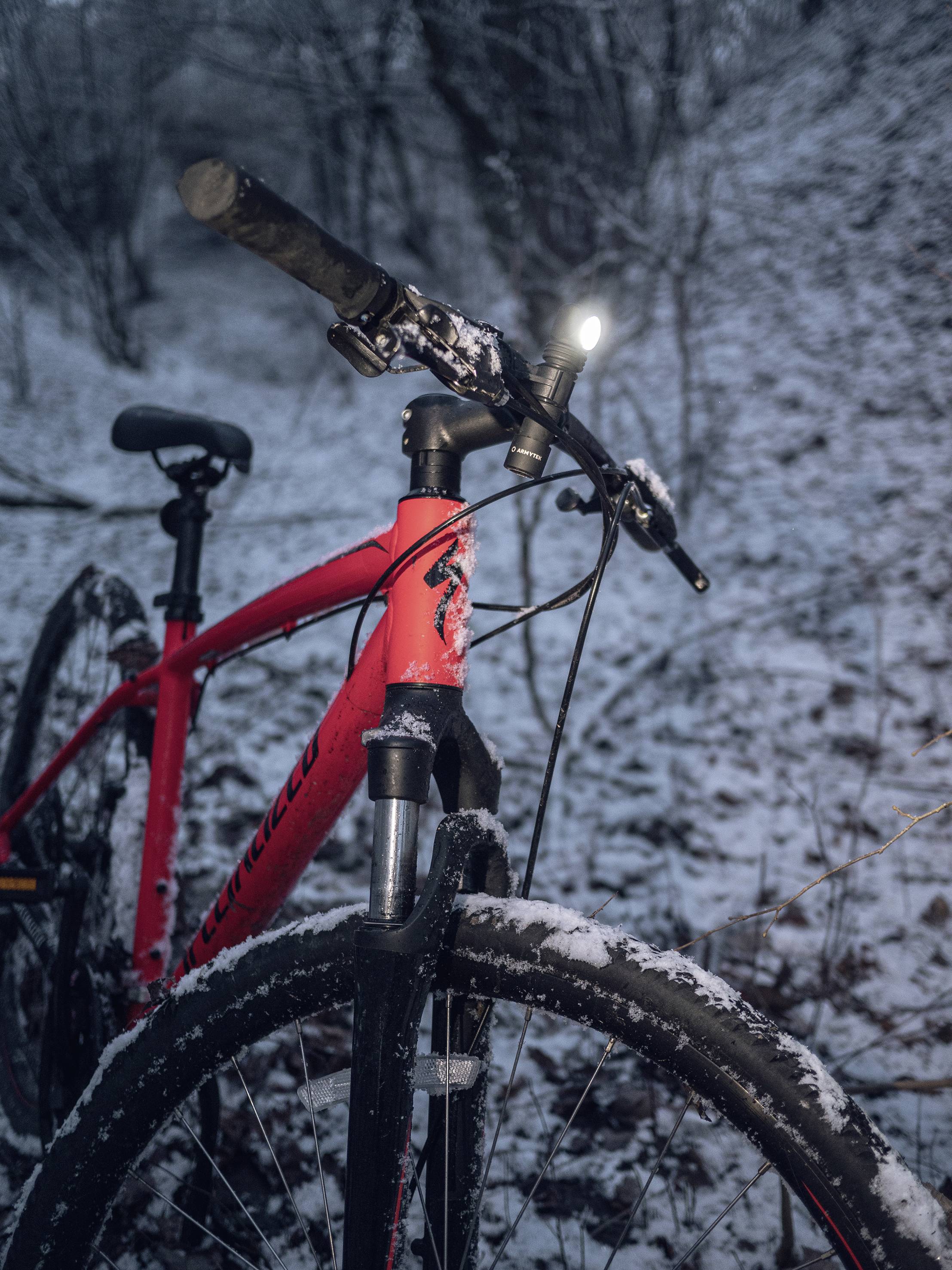 Ein rotes Fahrrad steht in einem verschneiten Waldweg. Das Vorderlicht leuchtet, um die Umgebung zu erhellen.