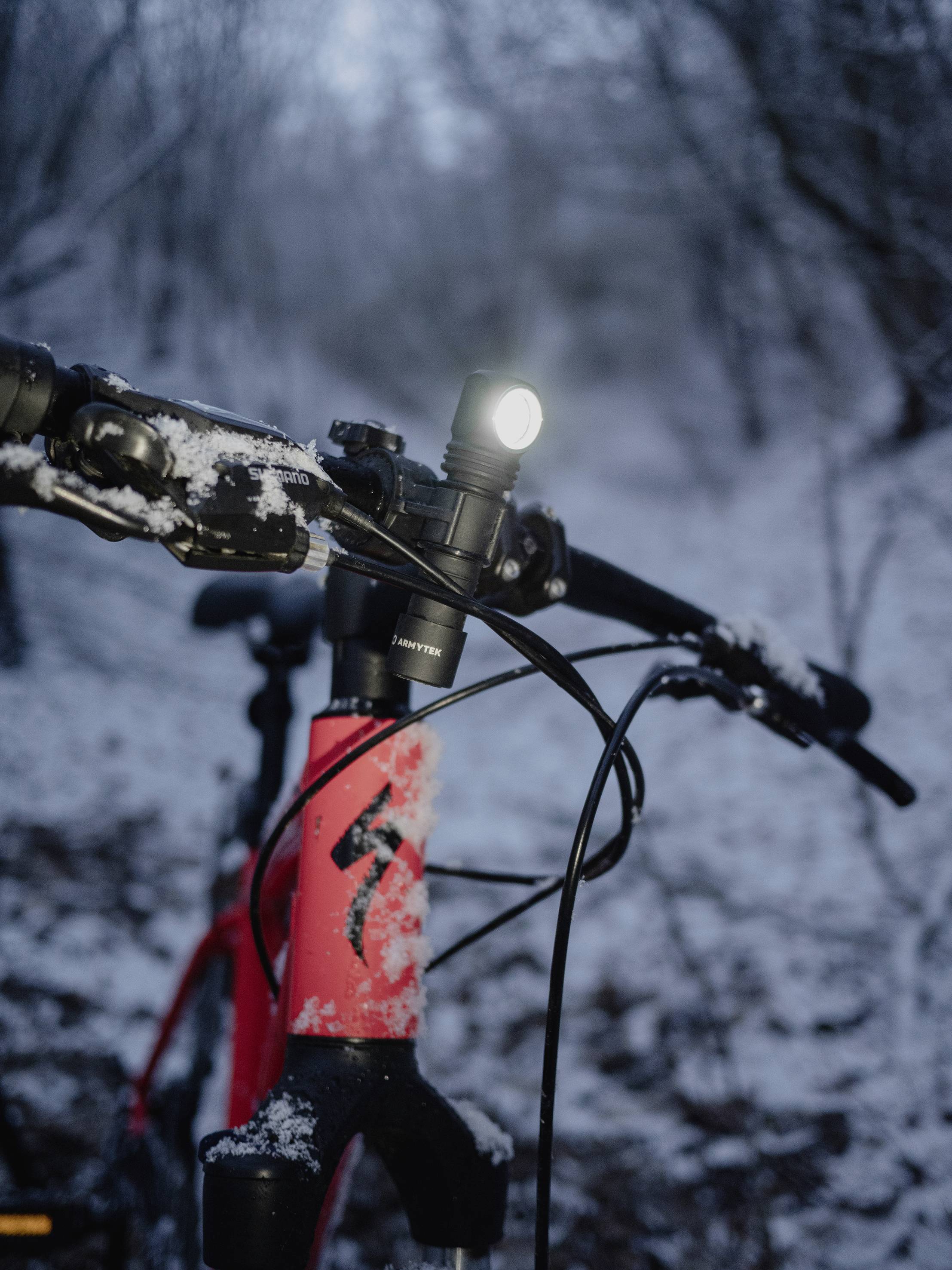 Nahaufnahme eines roten Fahrrads mit eingeschaltetem Vorderlicht, auf einem verschneiten Waldweg bei Dämmerung. Der Lenker ist teilweise mit Schnee bedeckt.