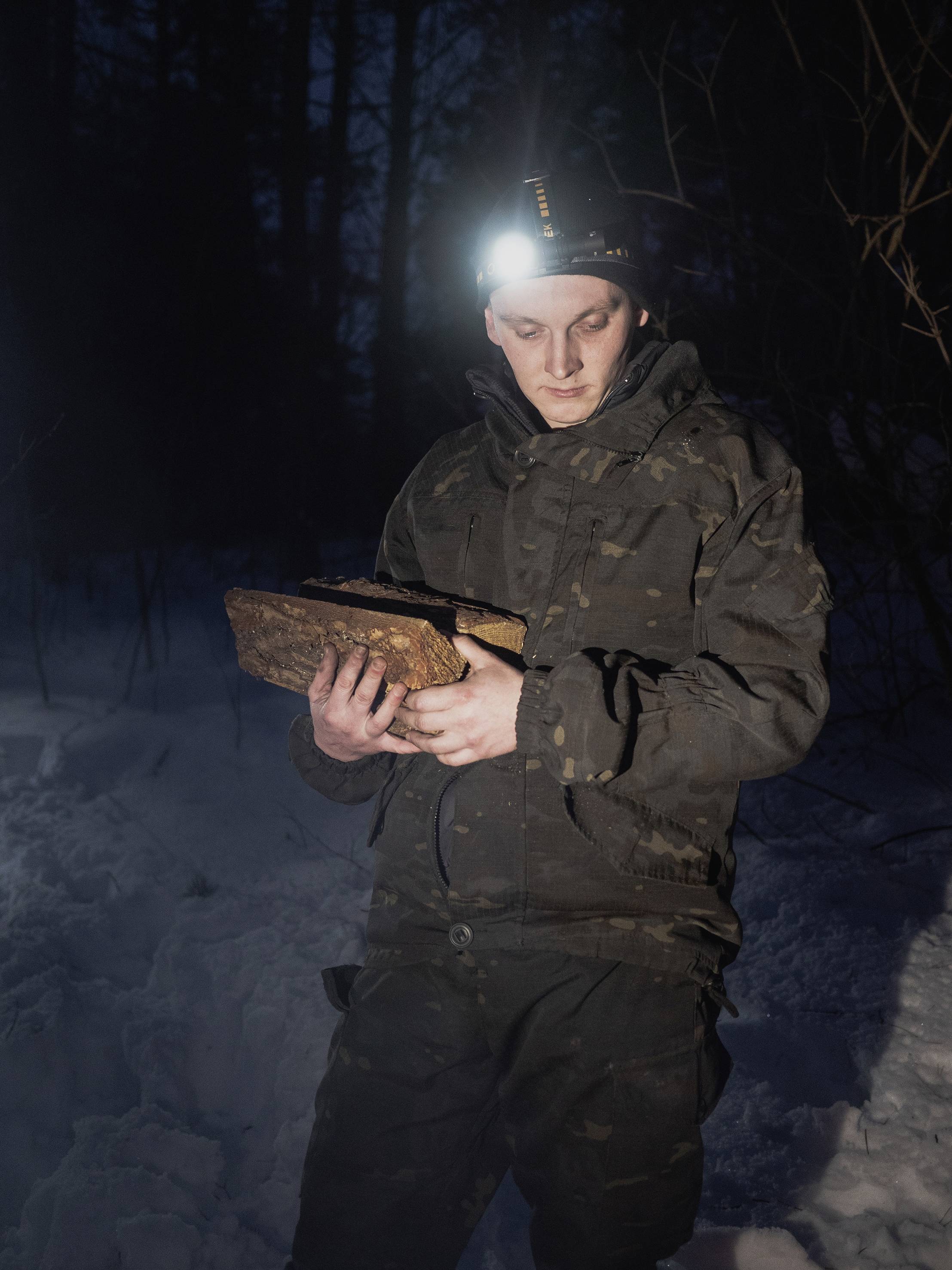 Ein Mensch in Tarnkleidung steht nachts im Schnee und hält ein rechteckiges Objekt aus Holz, beleuchtet von einer Stirnlampe.