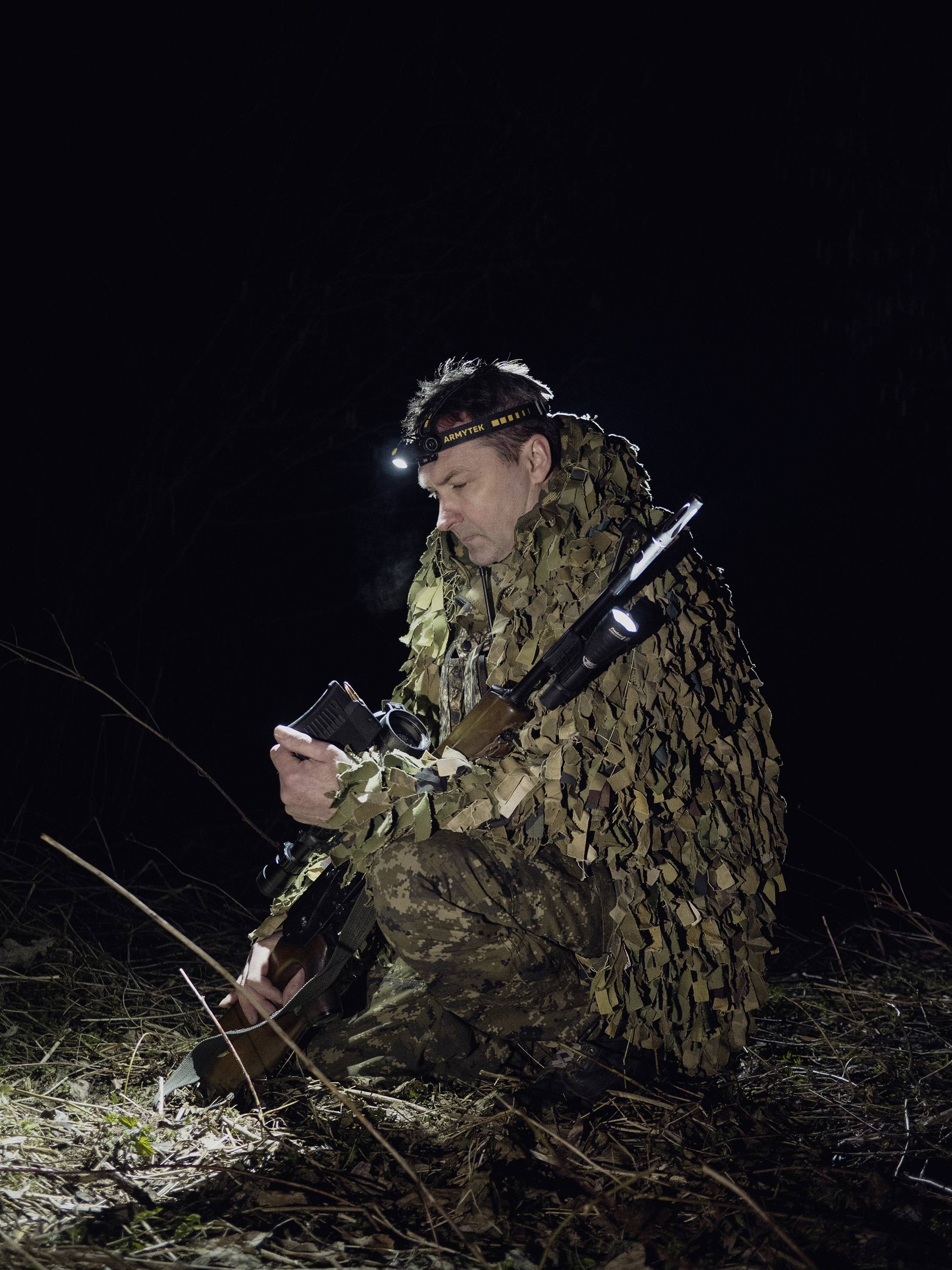 Eine Person in Tarnkleidung sitzt nachts im Wald und schaut auf ein Gerät in der Hand, trägt eine Kopflampe, neben sich ein Gewehr.
