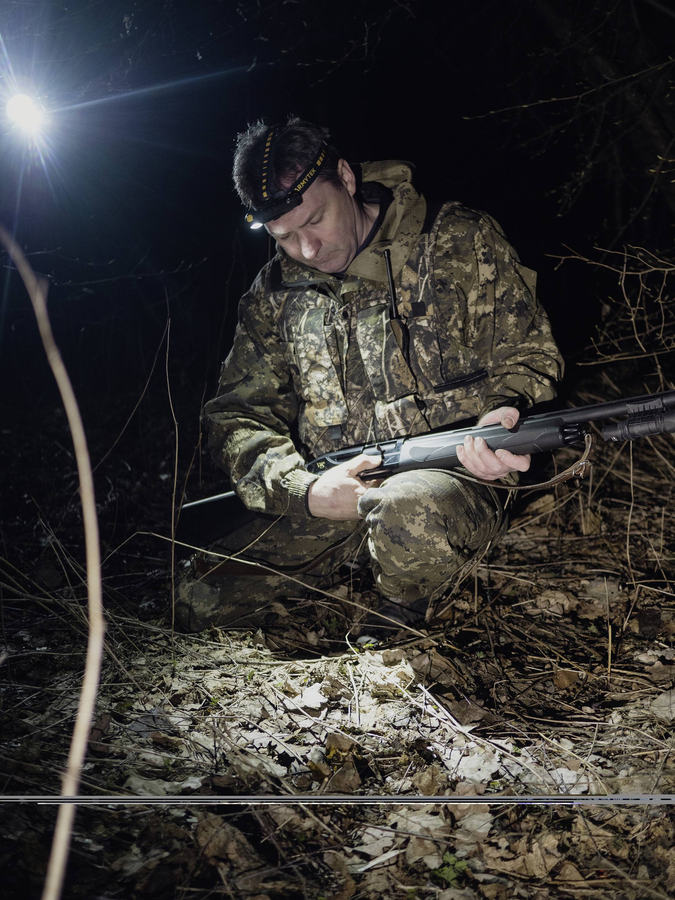 Eine Person in Tarnkleidung kniet im dunklen Wald, hält ein Gewehr und trägt eine Stirnlampe. Mögliches nächtliches Wildtierbeobachten.