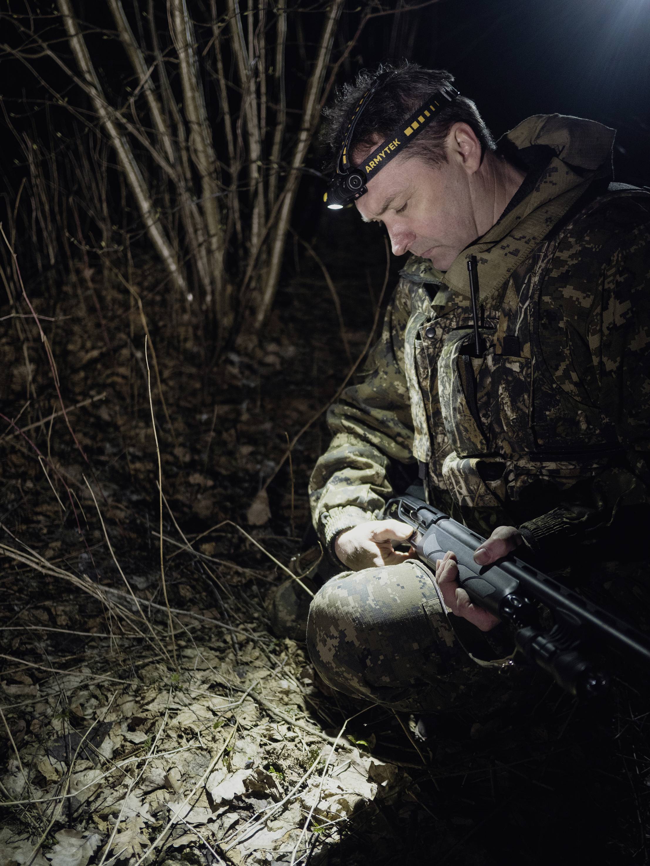 Ein Mann in Tarnkleidung kniet nachts im Wald und hält ein Gewehr. Eine Stirnlampe beleuchtet den Boden und einige trockene Äste.