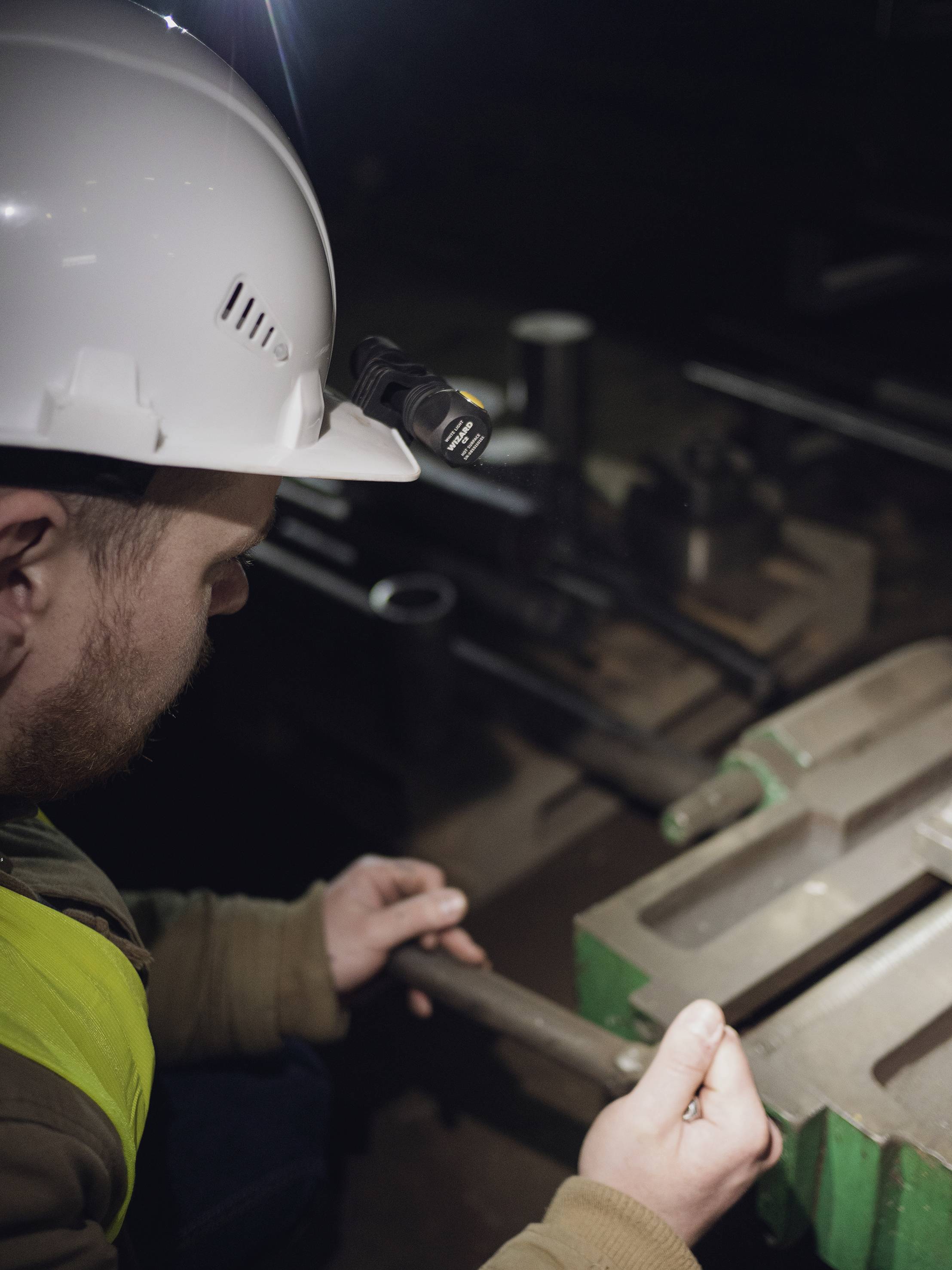Ein Arbeiter mit Helm und Stirnlampe bearbeitet ein Werkstück in einer dunklen Werkstatt, konzentriert auf eine präzise Aufgabe.