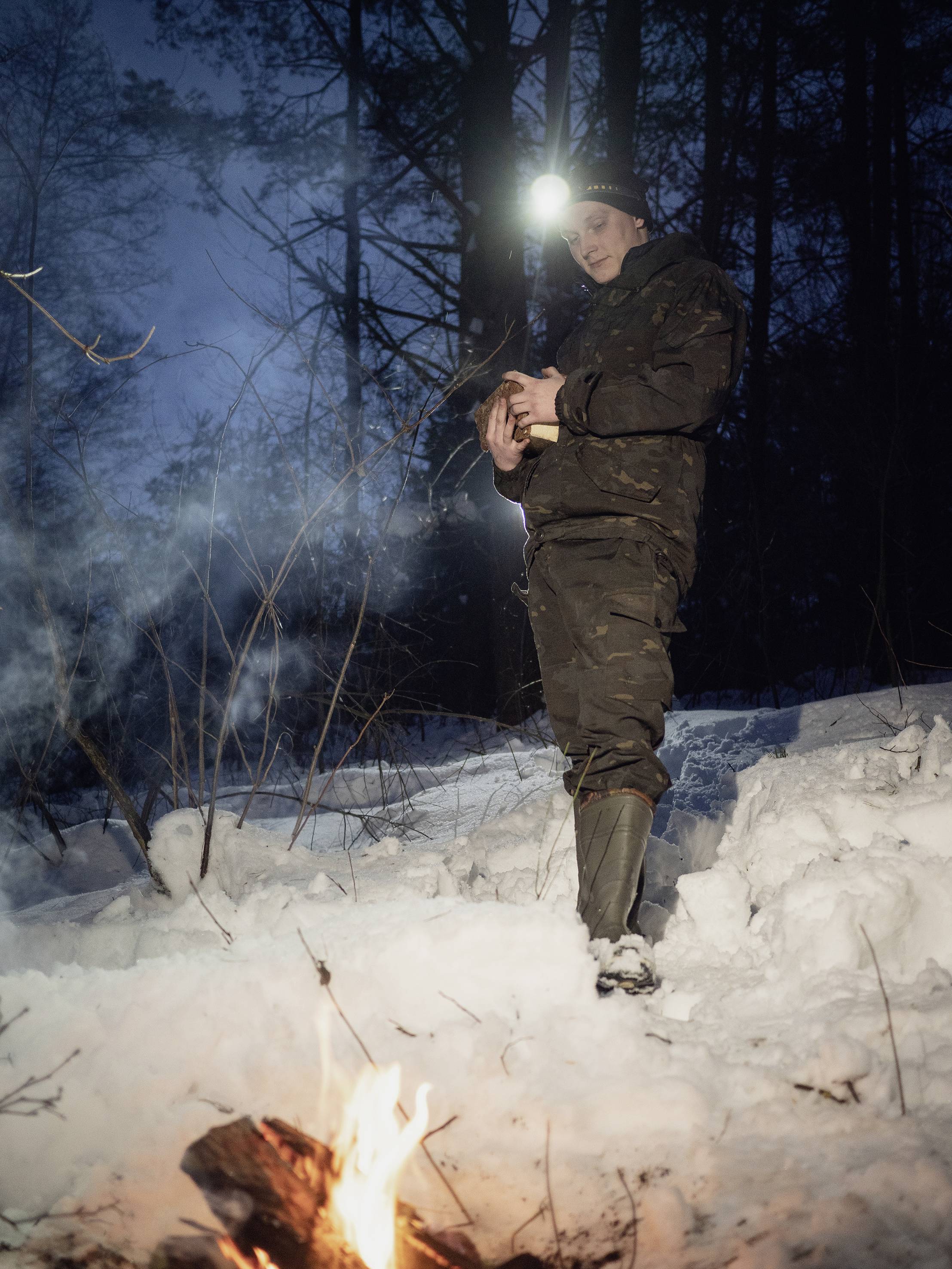 Person in Winterkleidung steht im Schnee im Wald, hält ein Tier im Arm. Im Vordergrund brennt ein kleines Lagerfeuer.