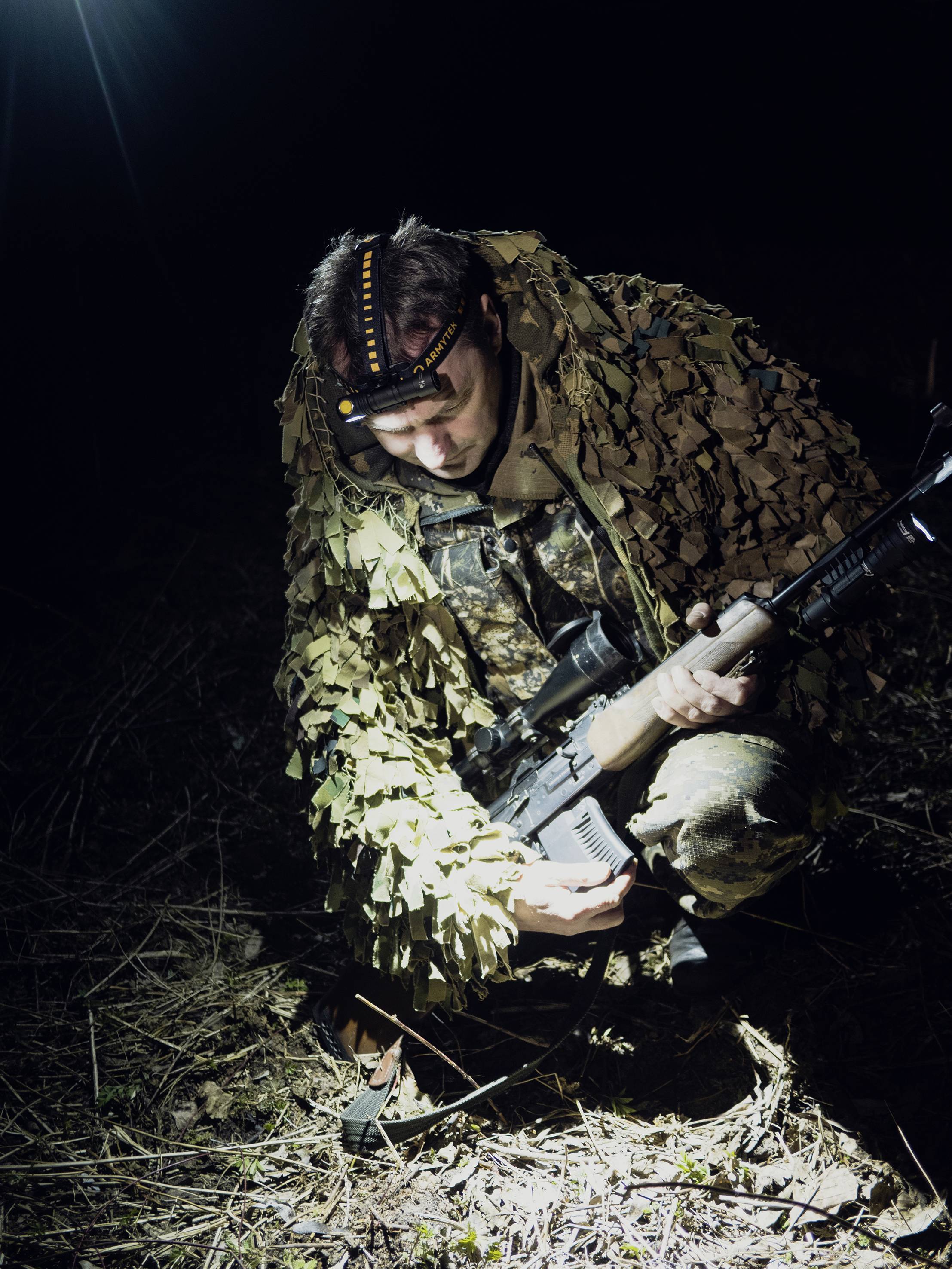 Ein Mensch in Tarnkleidung kniet nachts auf dem Boden, hält ein Gewehr in beiden Händen und wird von oben beleuchtet.