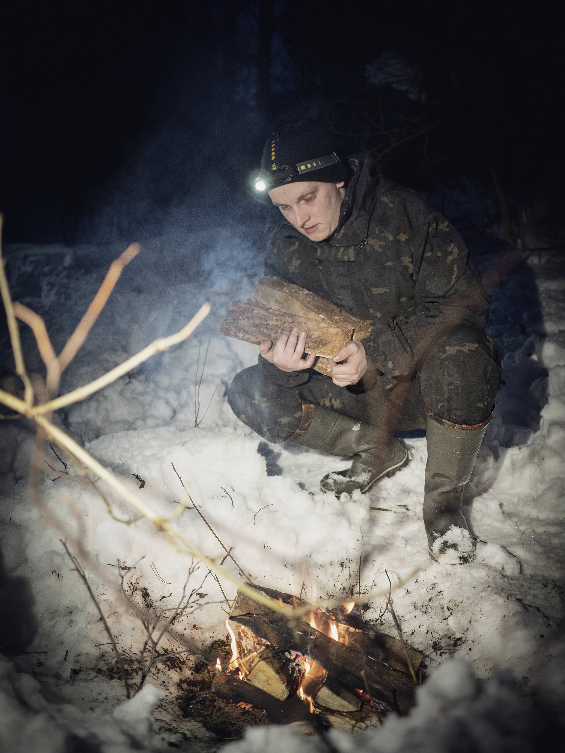 Eine Person im Wald legt Holzscheite auf ein Lagerfeuer im Schnee. Sie trägt eine Stirnlampe und warme Kleidung.