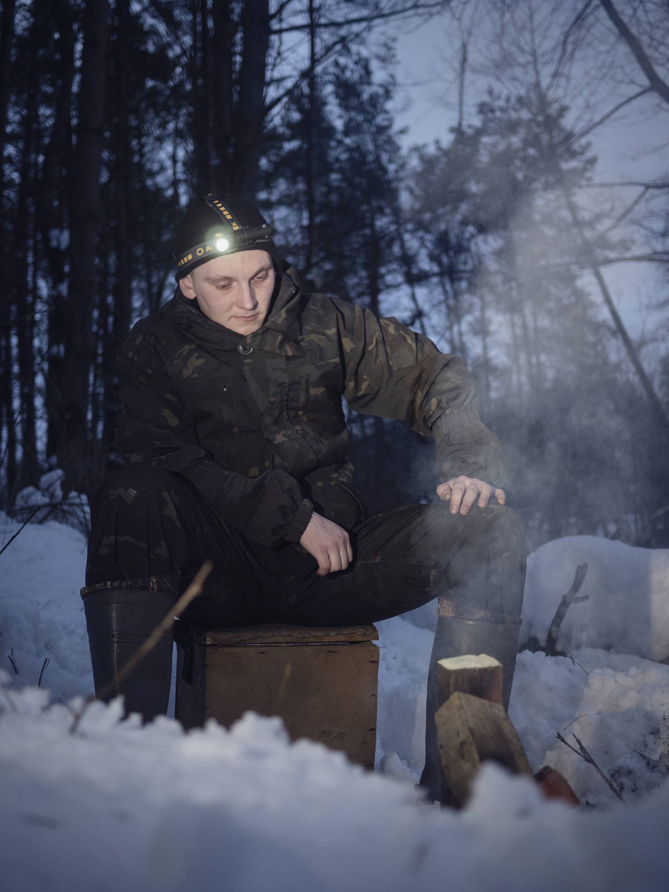 Person sitzt in dunkler Winterkleidung und Stirnlampe auf einem Holzkasten im Wald, umgeben von Schnee. Rauch steigt von einem kleinen Feuer auf.