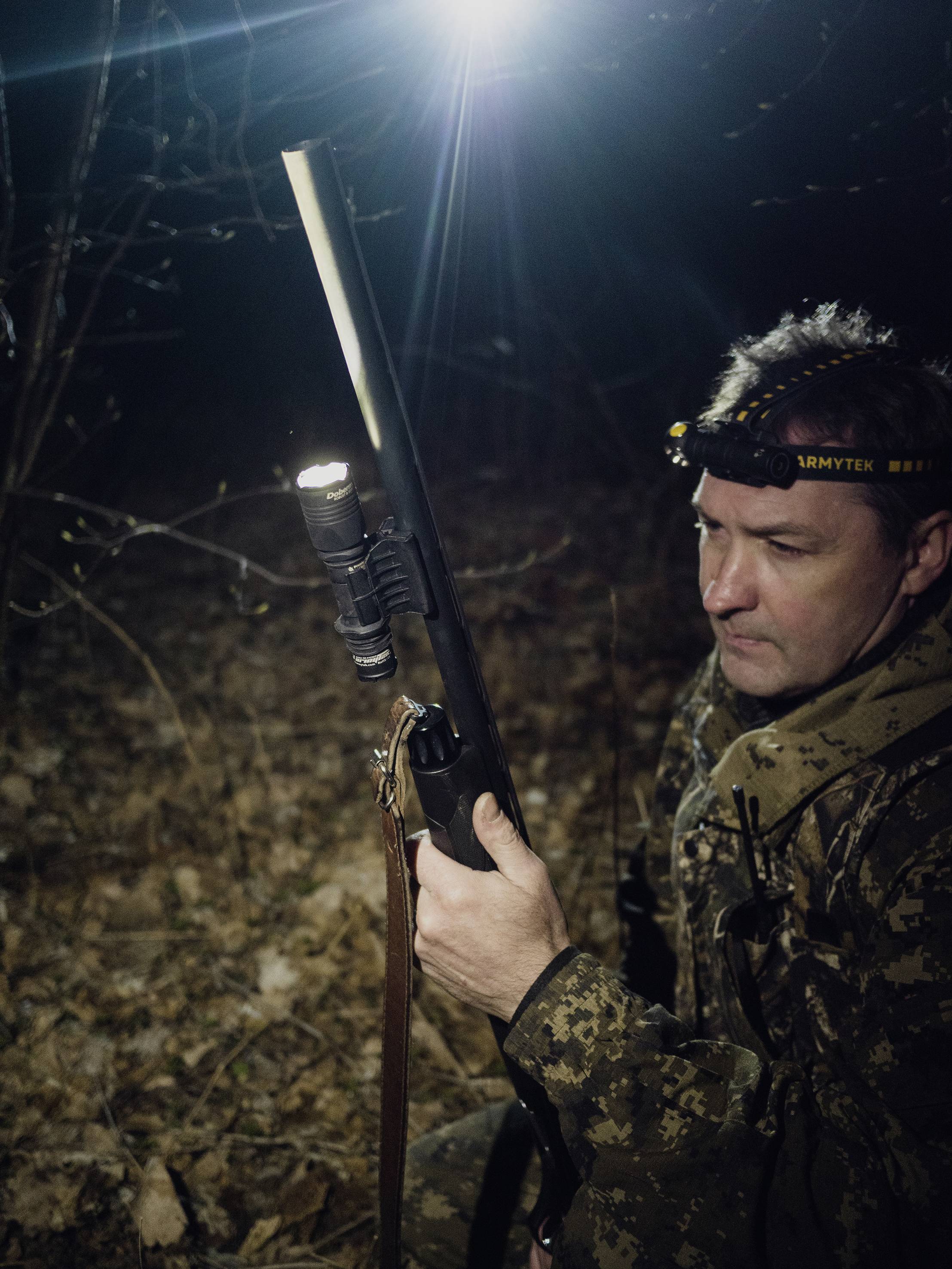 Eine Person in Tarnkleidung hält nachts eine Jagdwaffe mit Taschenlampe, umgeben von Wald. Eine Stirnlampe leuchtet den Weg.
