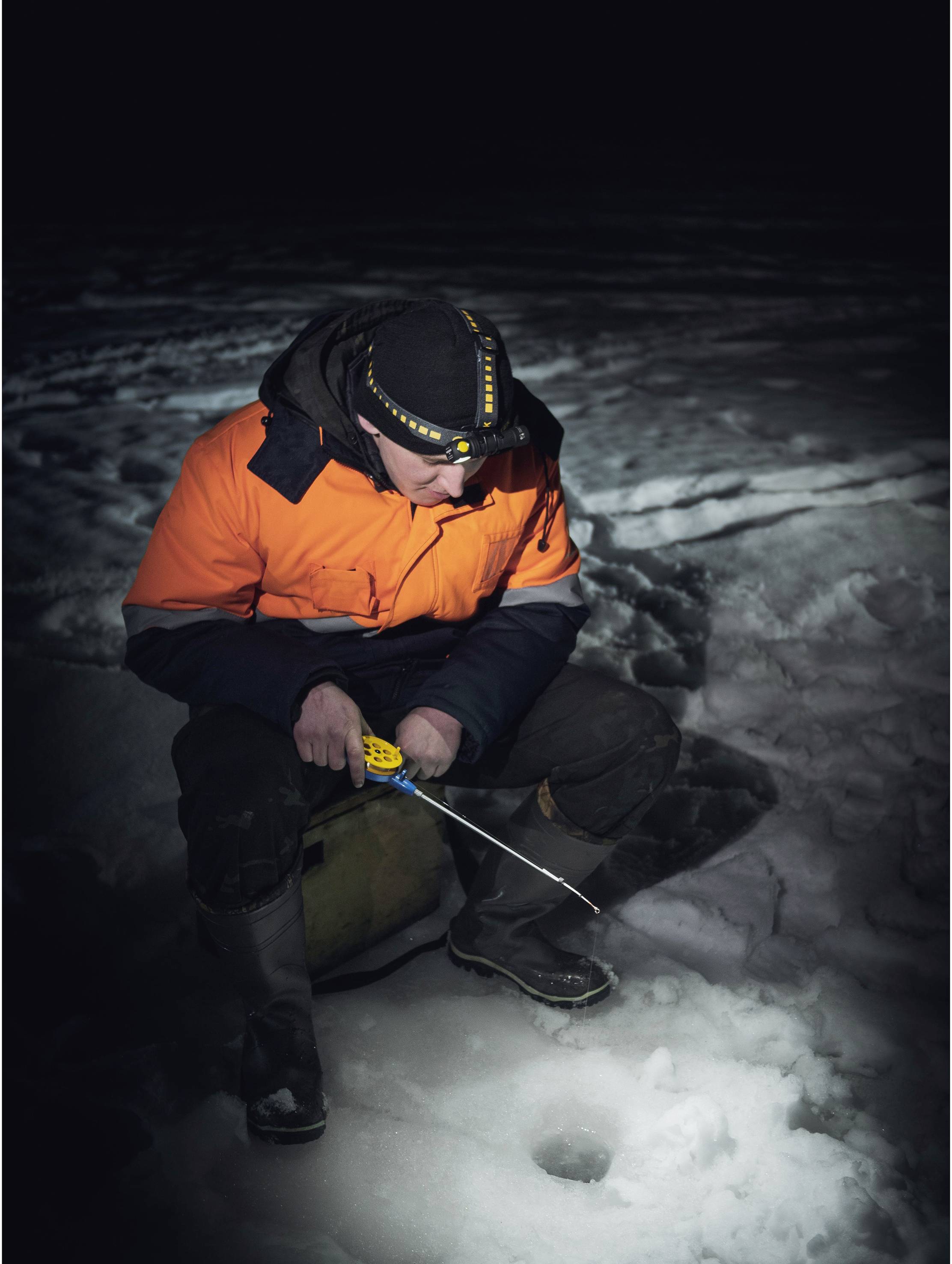 Eine Person in einer leuchtend orangefarbenen Jacke sitzt auf einem gefrorenen See, beim Eisfischen durch ein Loch im Eis. Dunkle Nachtumgebung.
