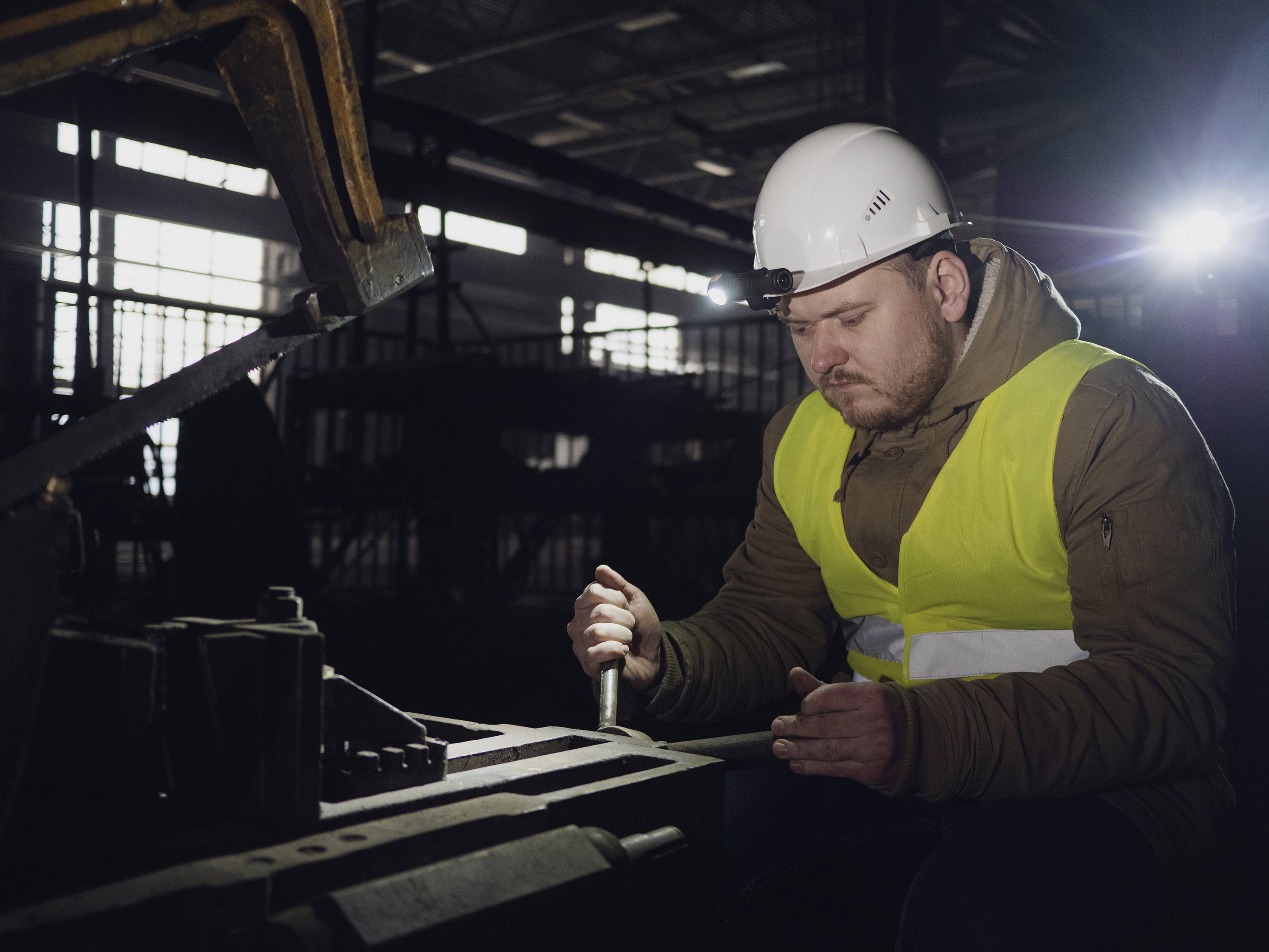 Ein Arbeiter in Sicherheitskleidung bearbeitet mit einem Werkzeug ein Metallstück in einer Fabrikhalle; gedämpftes Licht im Hintergrund.
