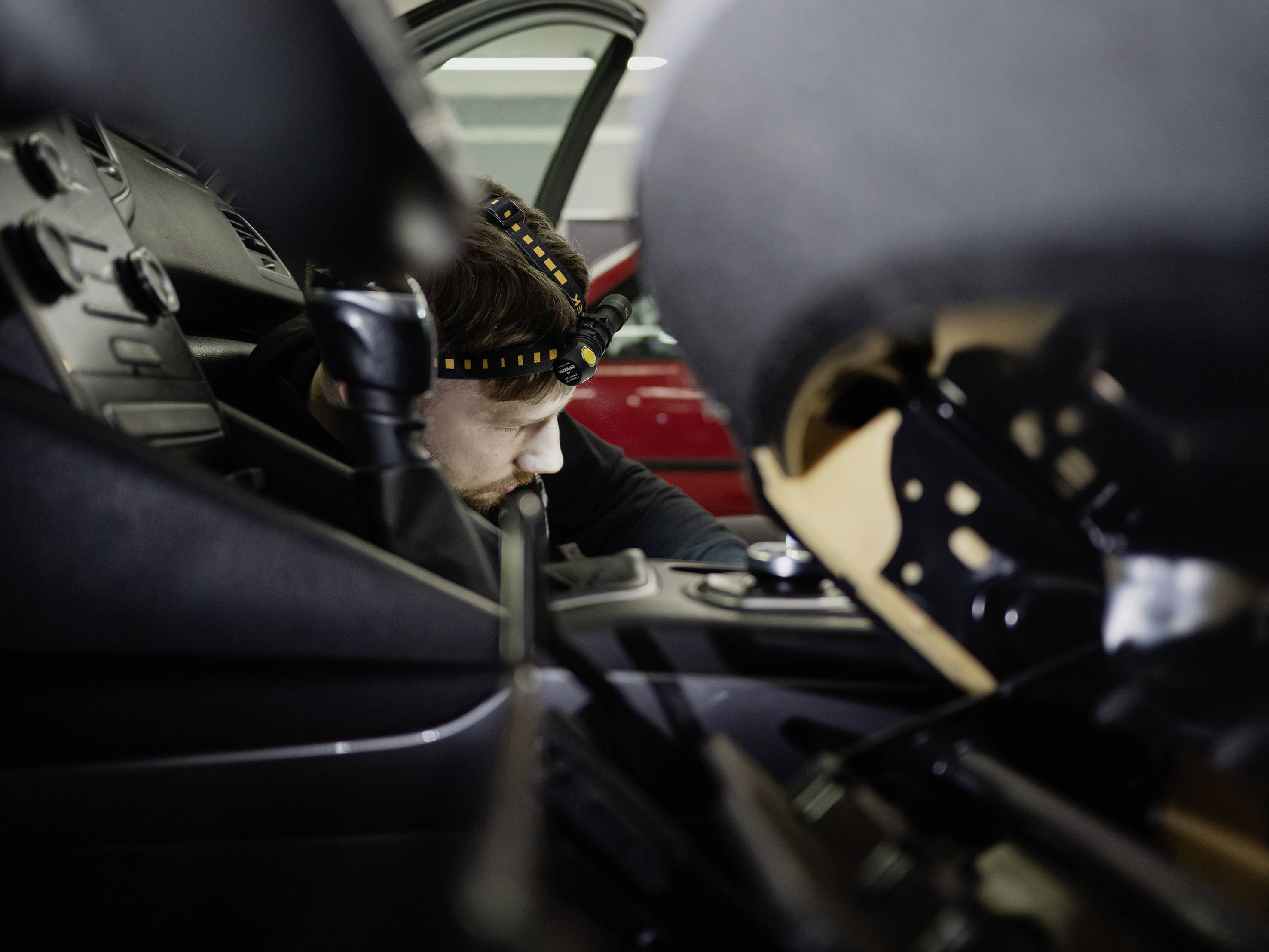 Person arbeitet an der Innenausstattung eines Autos mit einem Stirnlicht. Ein rotes Auto ist im Hintergrund teilweise sichtbar.