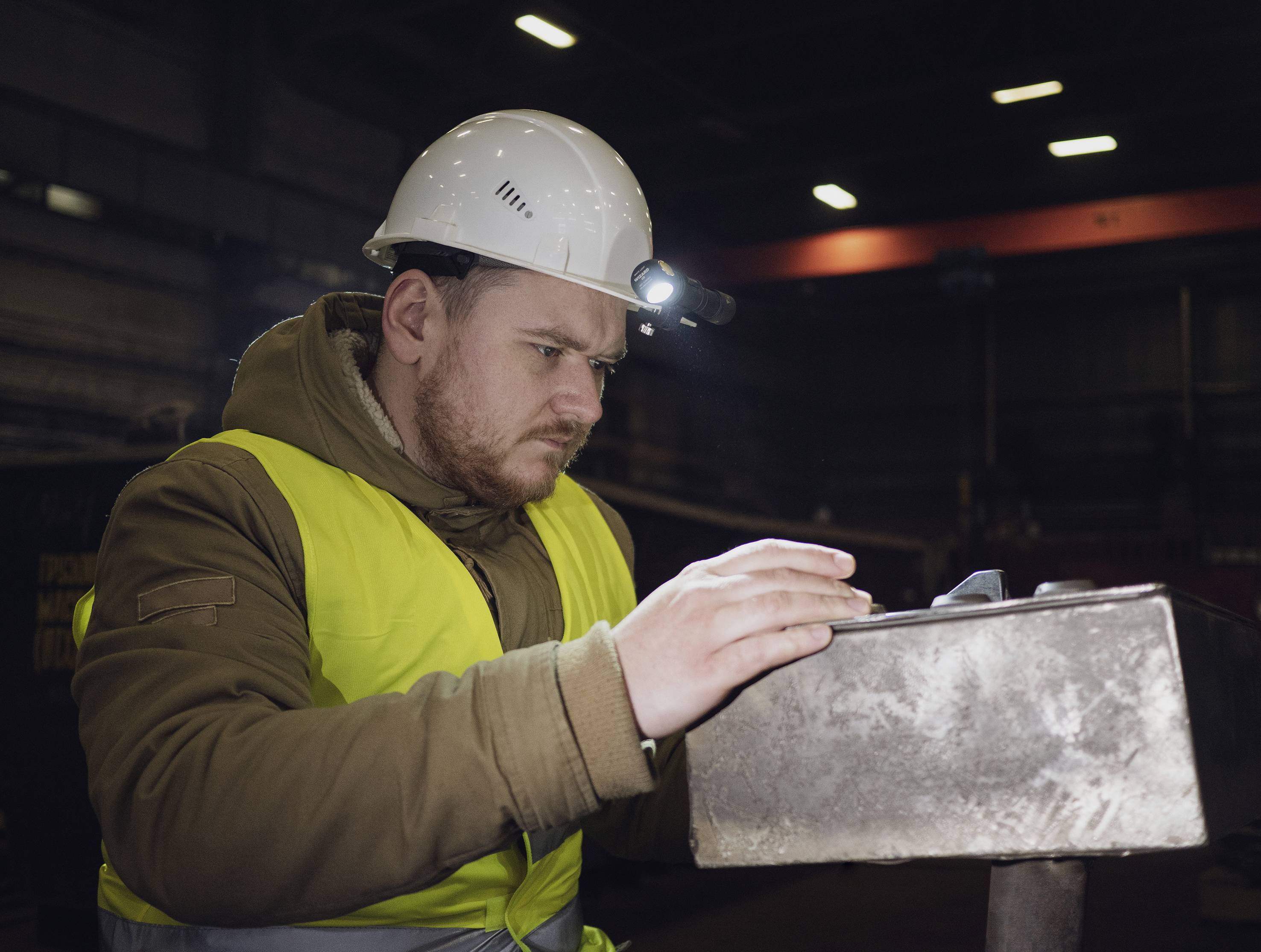Ein Mann in einem gelben Sicherheitswesten und Helm bei der Arbeit in einer Fabrikhalle. Er konzentriert sich auf ein Metallteil.