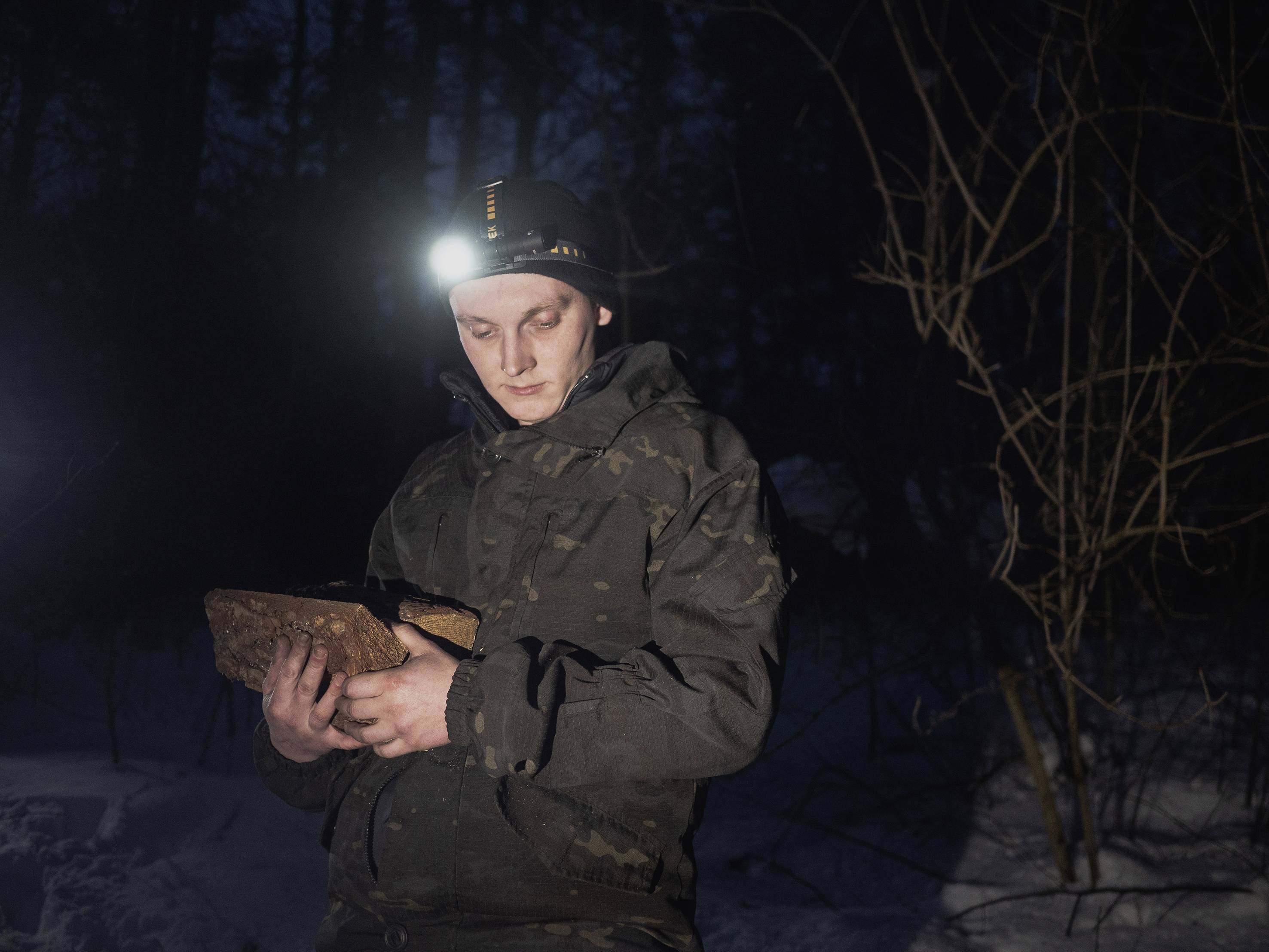 Eine Person im Tarnanzug steht nachts im Wald und liest ein Buch. Die Stirnlampe beleuchtet das Buch, während es dunkel um sie herum ist.