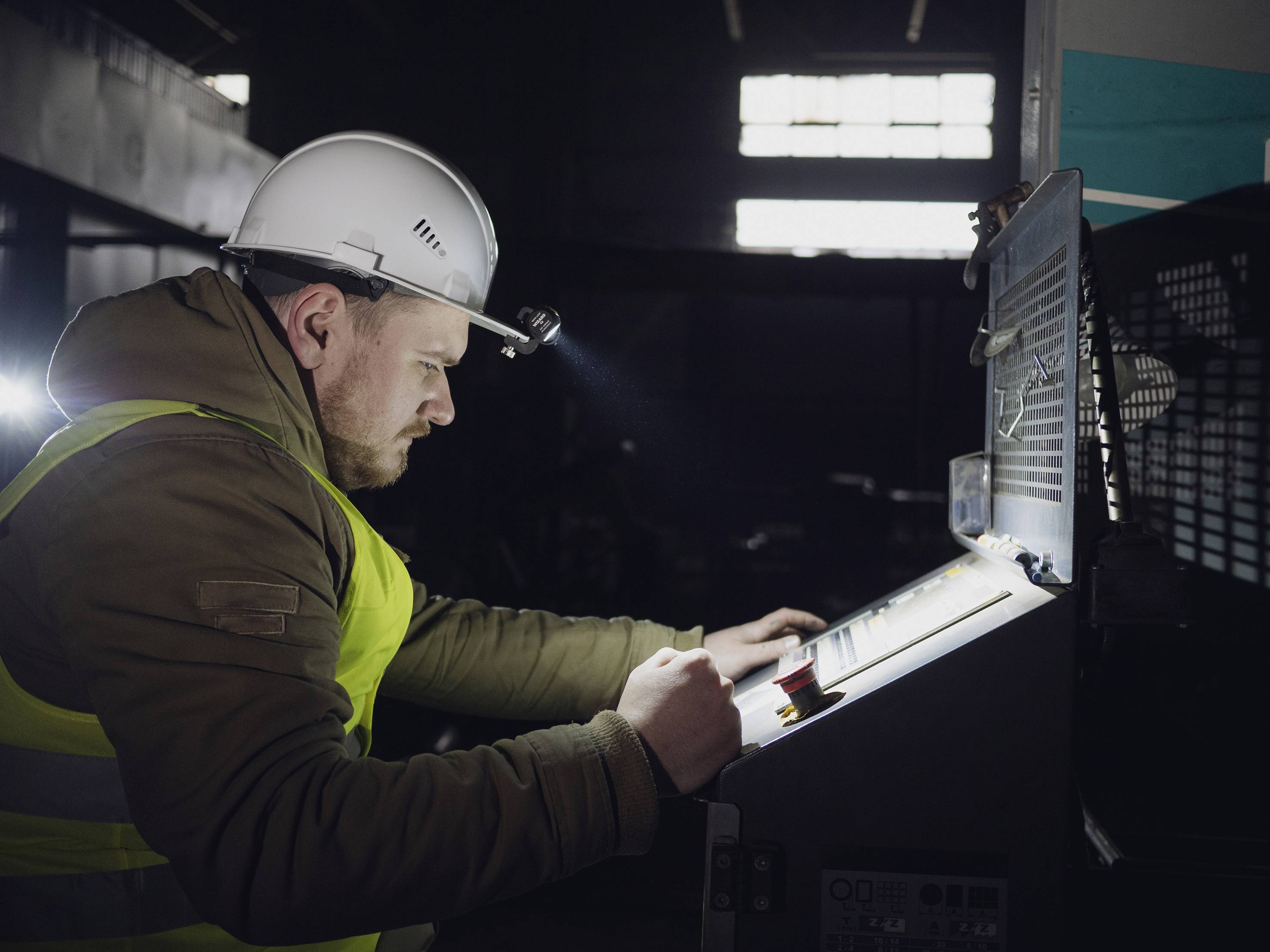 Arbeiter mit Helm und Schutzweste bedient in dunkler Umgebung eine industriell aussehende Maschine mit Kontrollbildschirm.