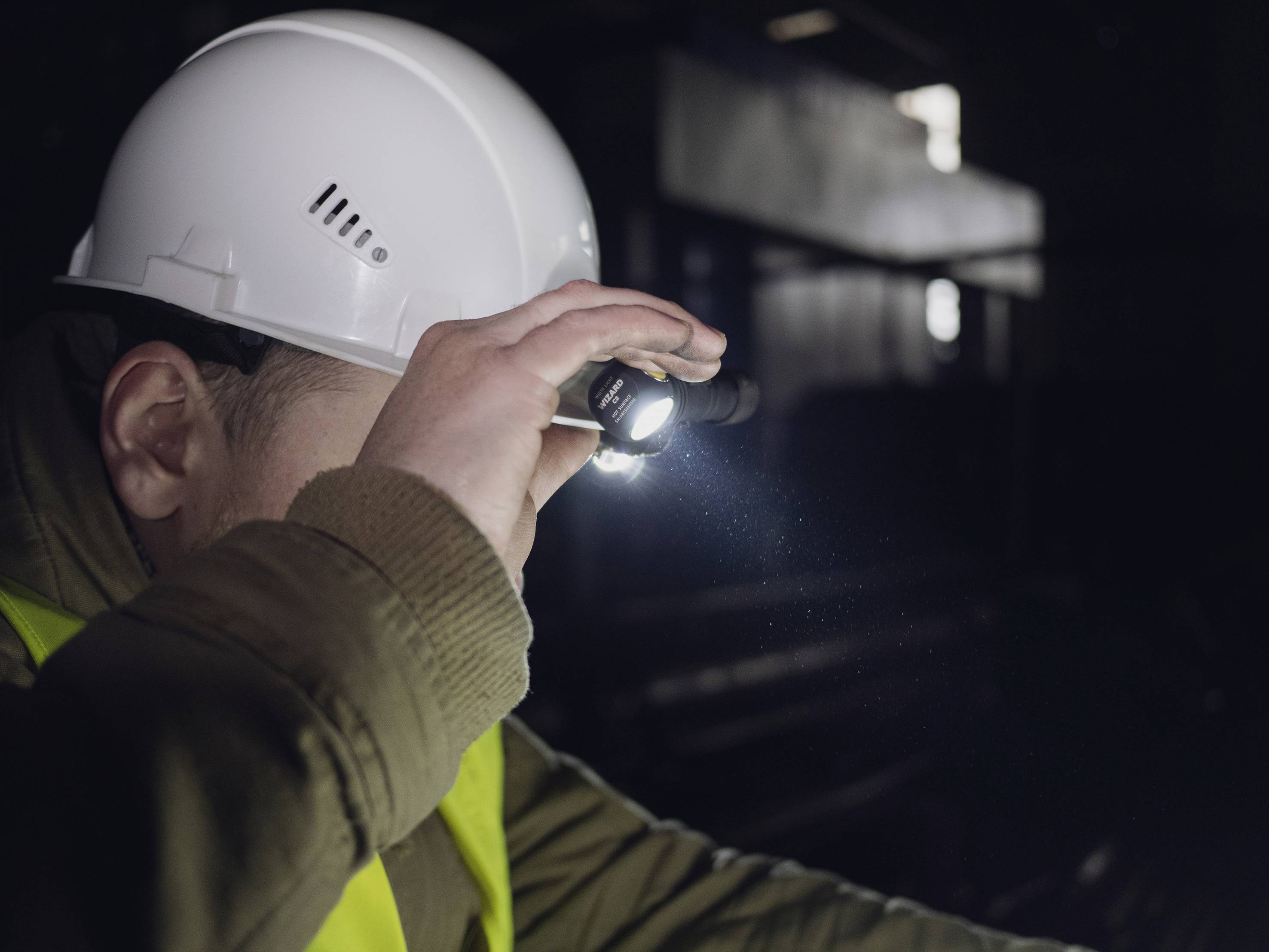 Eine Person in Arbeitskleidung und Helm untersucht einen dunklen Raum mit einer Taschenlampe, möglicherweise in einem industriellen Umfeld.
