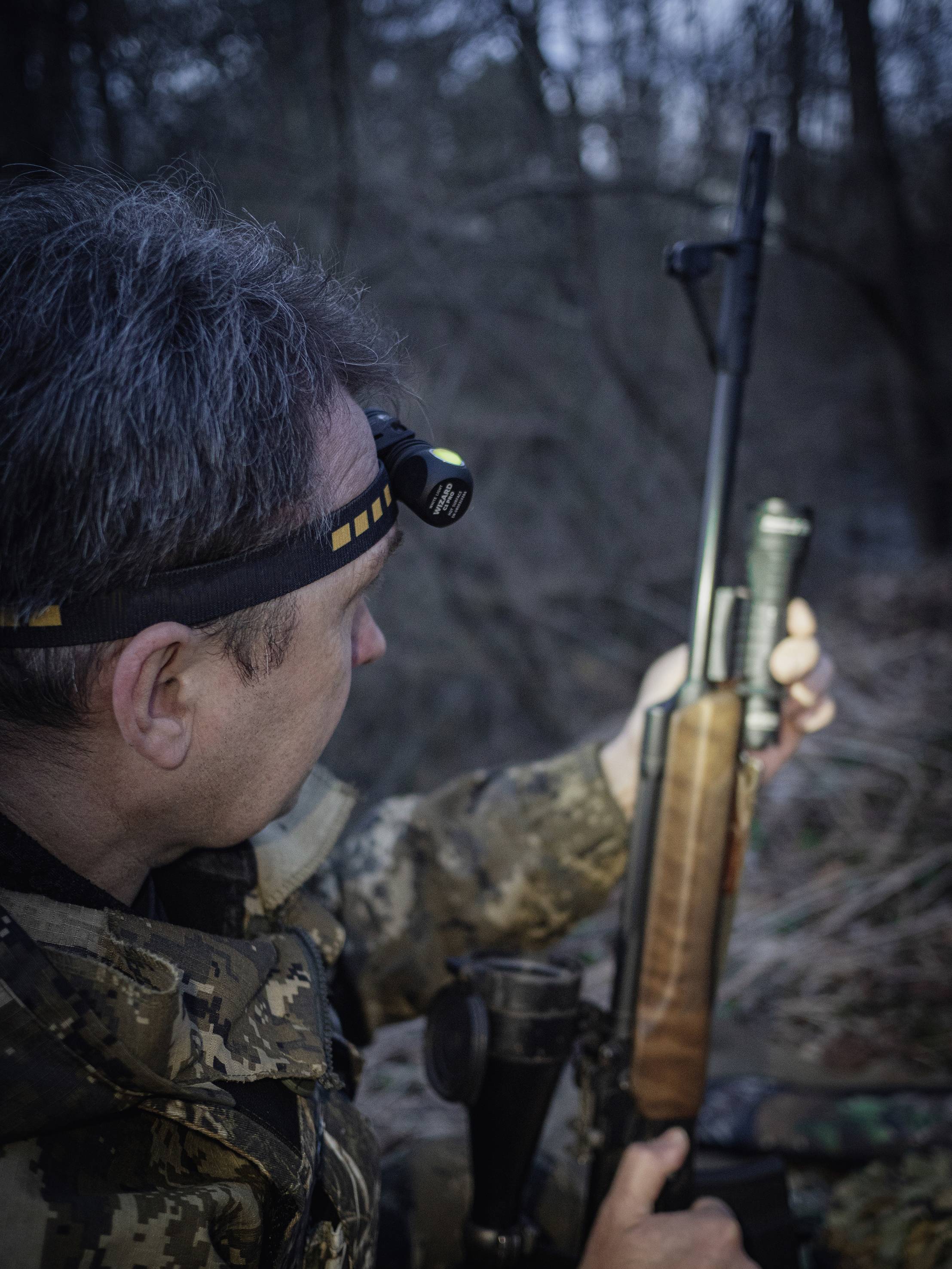 Eine Person im Tarnanzug hält ein Gewehr im Wald. Sie trägt eine Stirnlampe, die in der Dämmerung leuchtet, um die Umgebung zu erhellen.