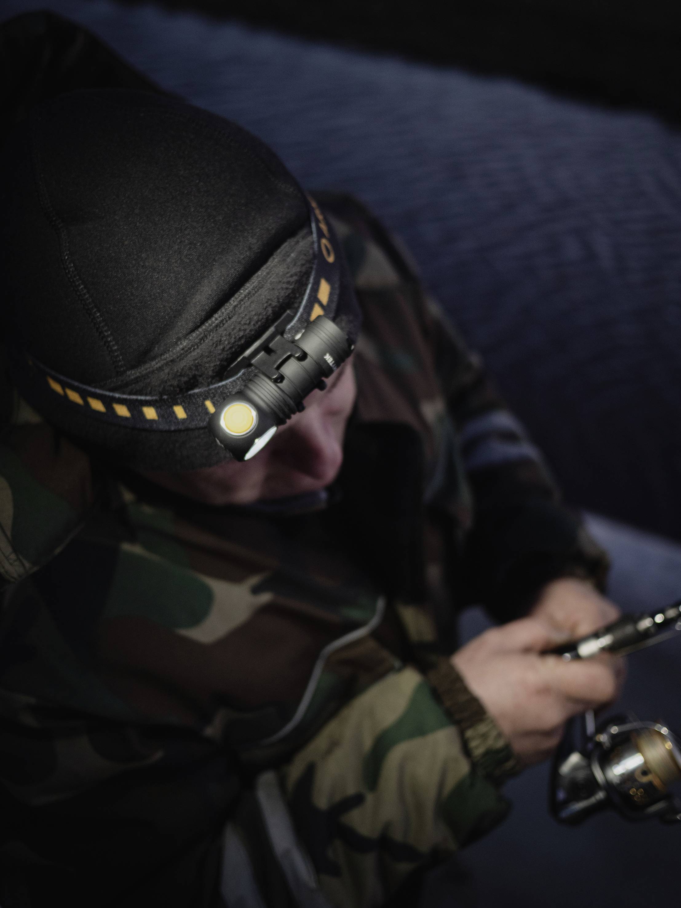 Eine Person in Tarnkleidung mit Stirnlampe beim Fischen hält eine Angelrute, konzentriert bei der Arbeit, mit dunkler Umgebung im Hintergrund.