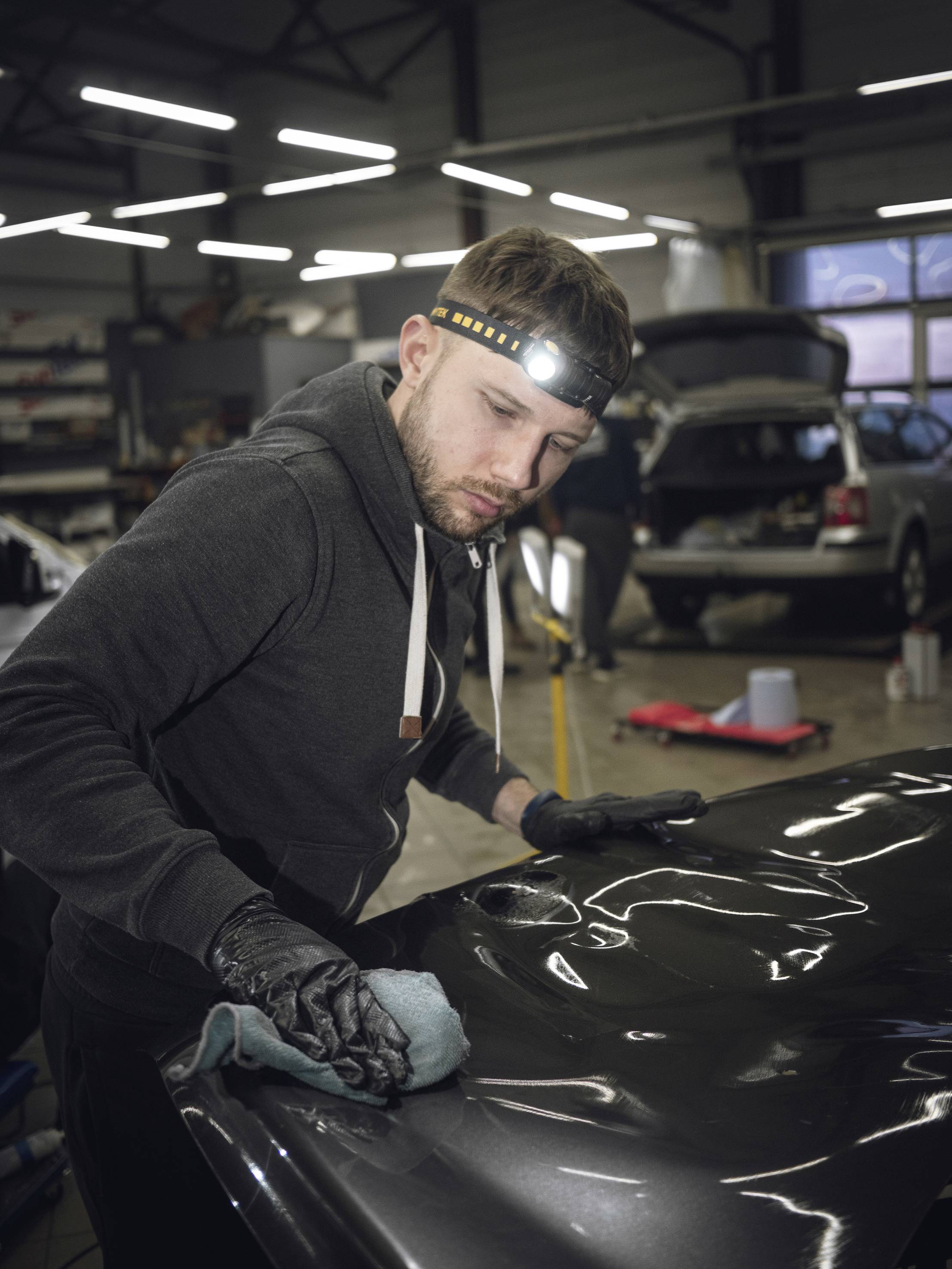Ein Mechaniker poliert die Motorhaube eines Autos in einer Werkstatt mit einer Stirnlampe auf dem Kopf, um die Lackierung zu verbessern.