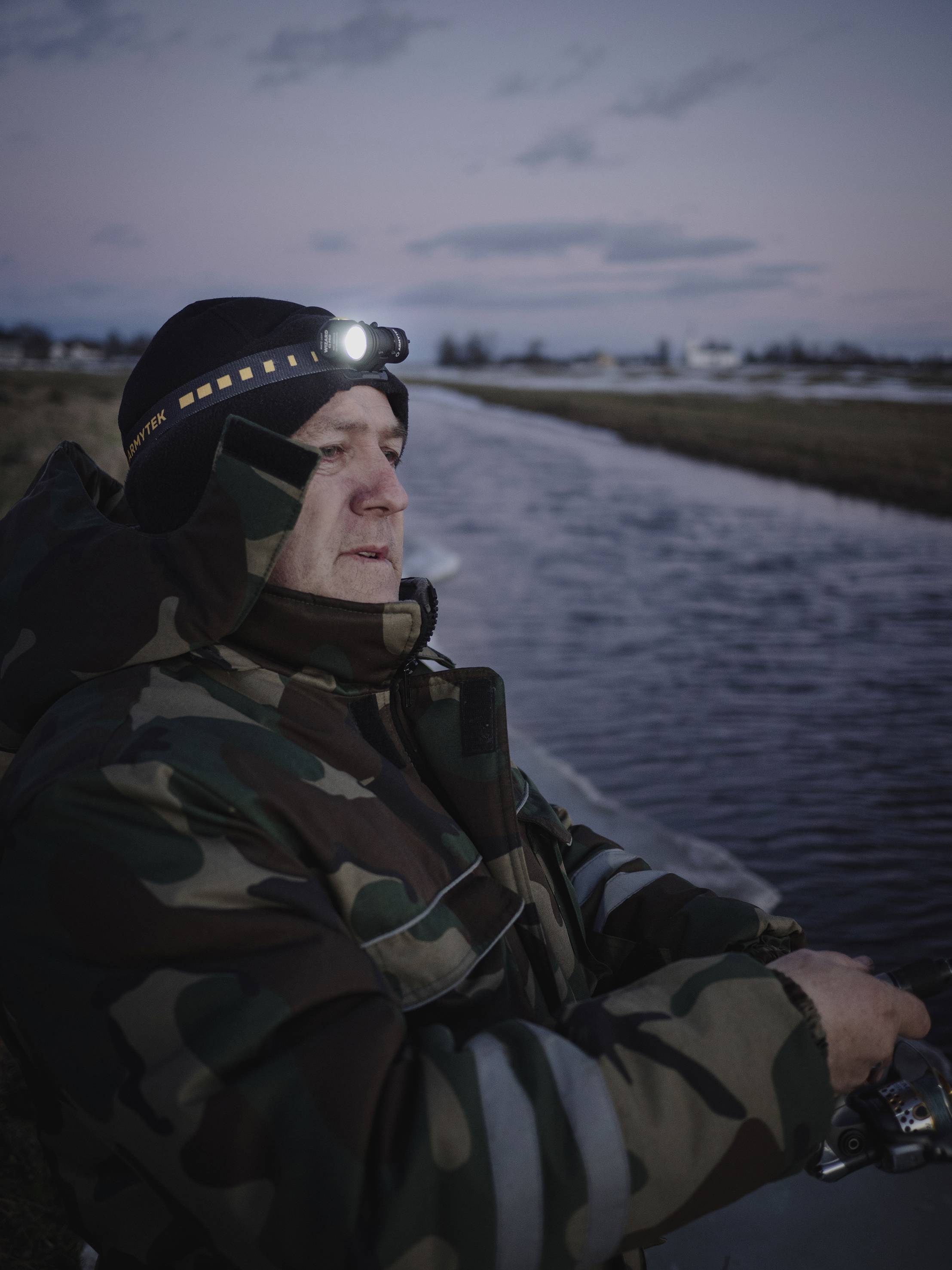 Ein Angler in Tarnkleidung steht an einem Fluss bei Dämmerung, trägt eine Stirnlampe und hält eine Angel in den Händen.