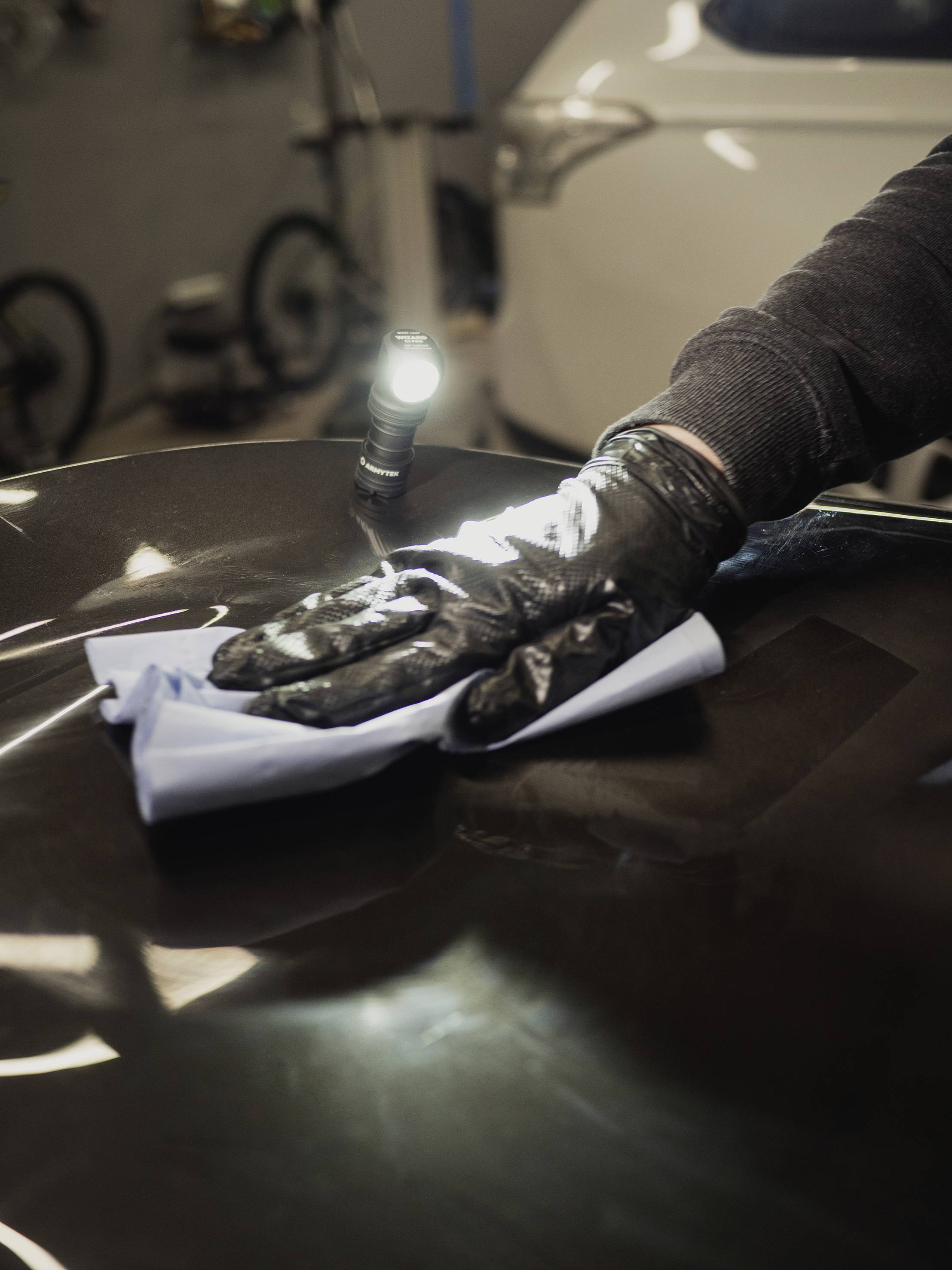 Eine Person reinigt mit einem Tuch und Handschuhen die Motorhaube eines Autos, beleuchtet von einer Lampe. Im Hintergrund sind Fahrräder sichtbar.
