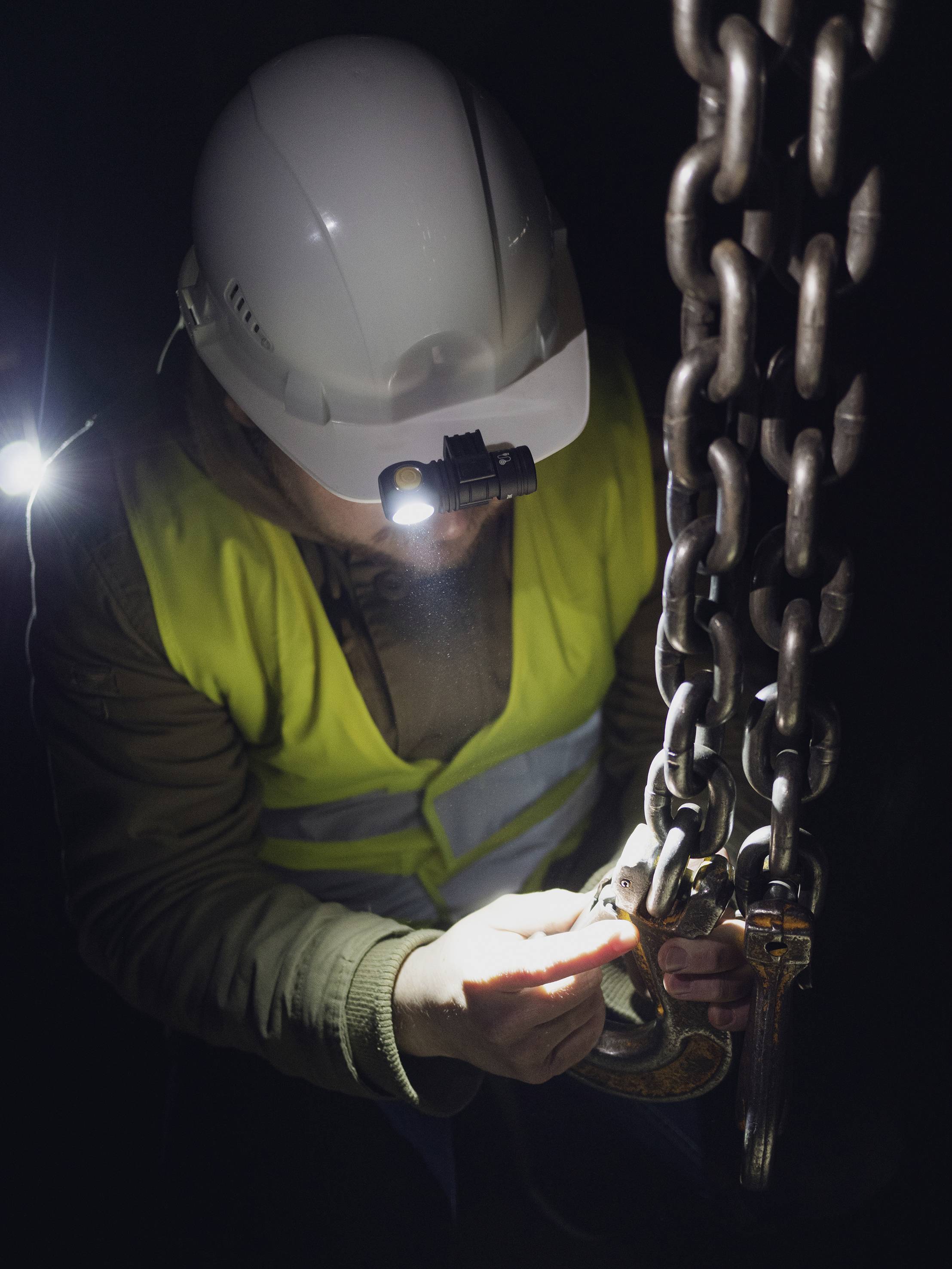 Arbeiter mit Helm und Stirnlampe inspiziert Kettenglied in dunkler Umgebung. Sicherheit und Präzision stehen im Mittelpunkt.