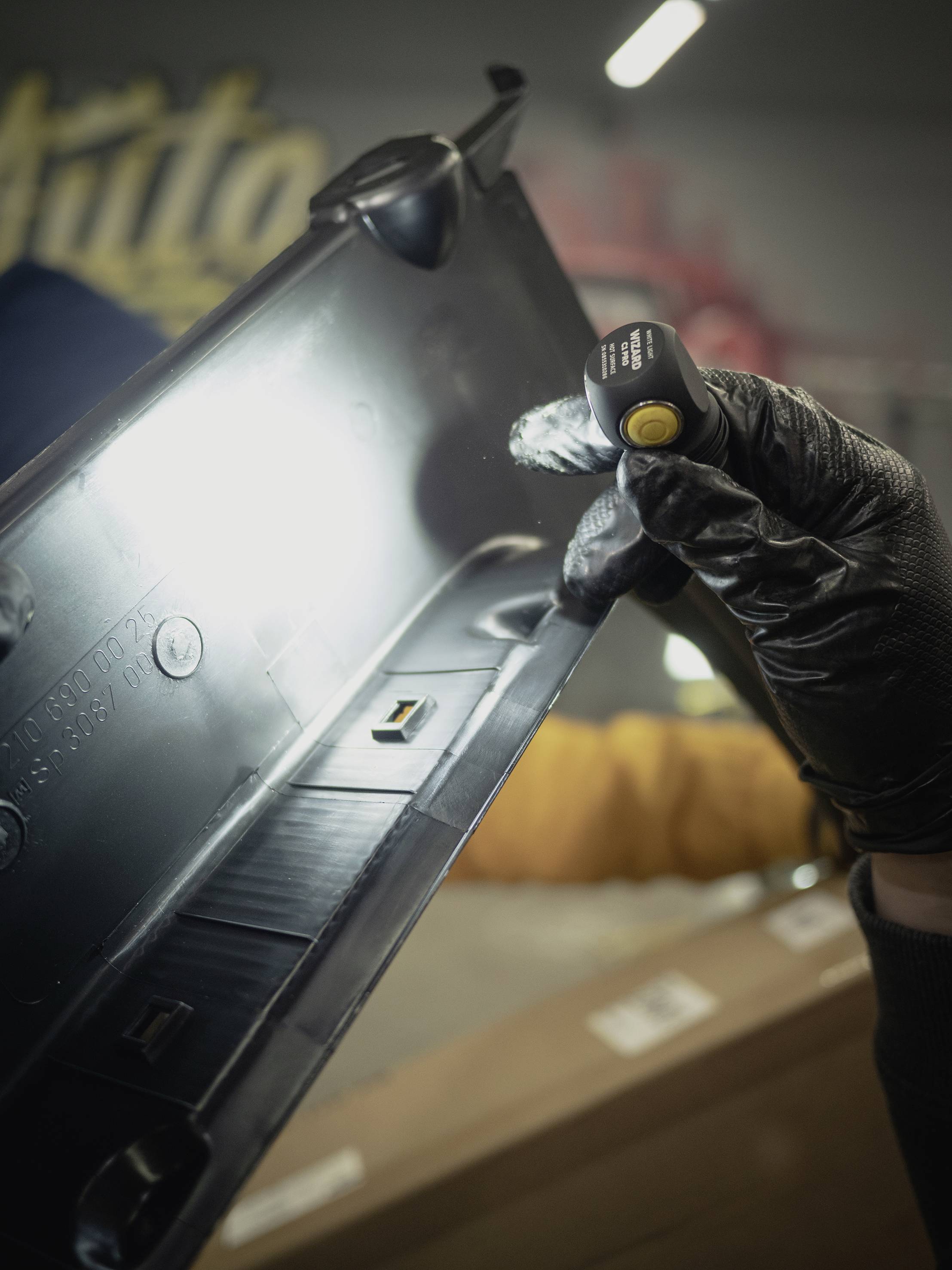 Eine Person mit schwarzen Handschuhen hält ein schwarzes Plastikteil gegen das Licht, um es zu inspizieren.