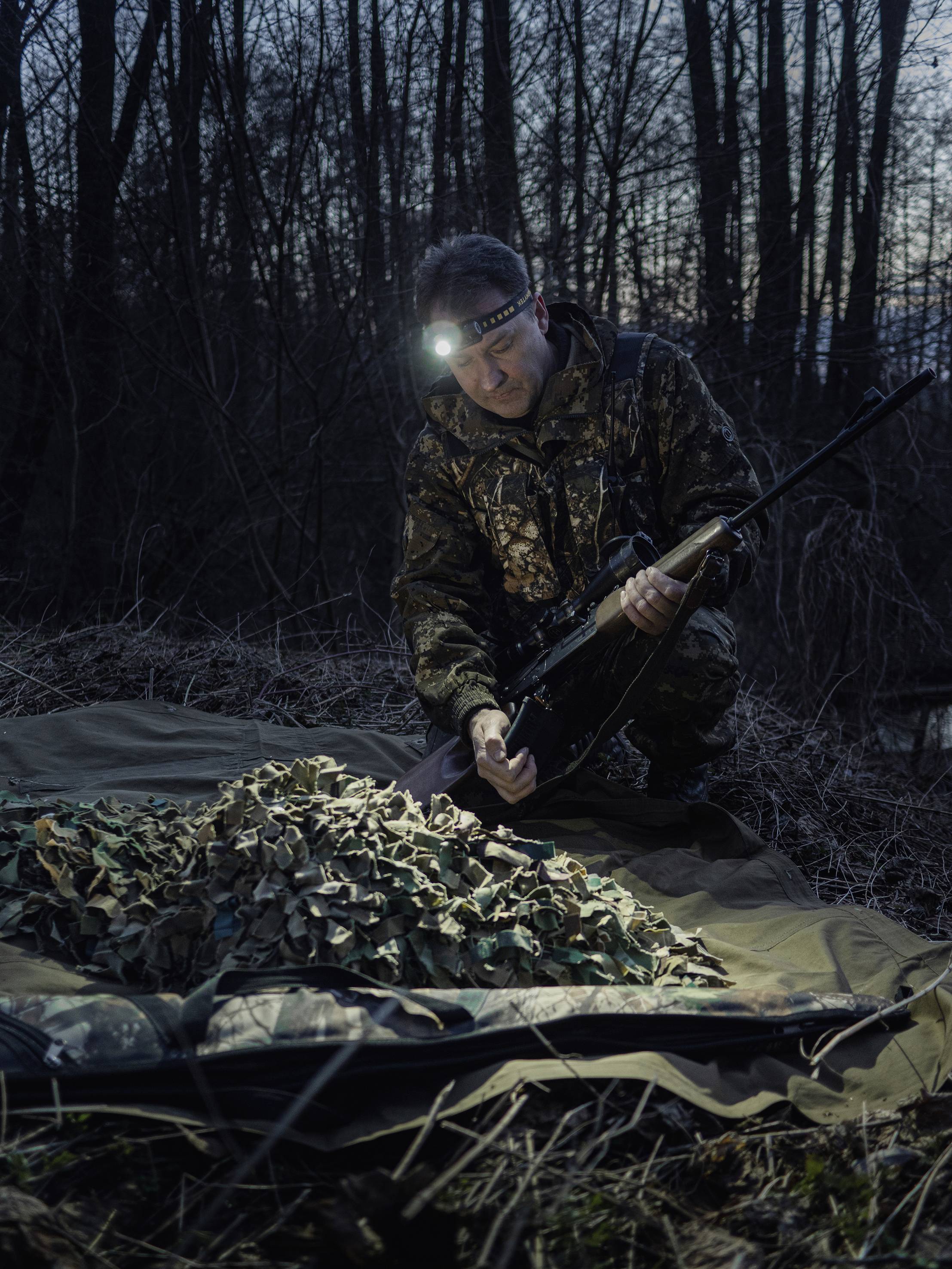 Ein Mann in Tarnkleidung und Stirnlampe hält ein Gewehr und kniet bei einem Haufen Tarnmaterial im Wald bei Dämmerung.