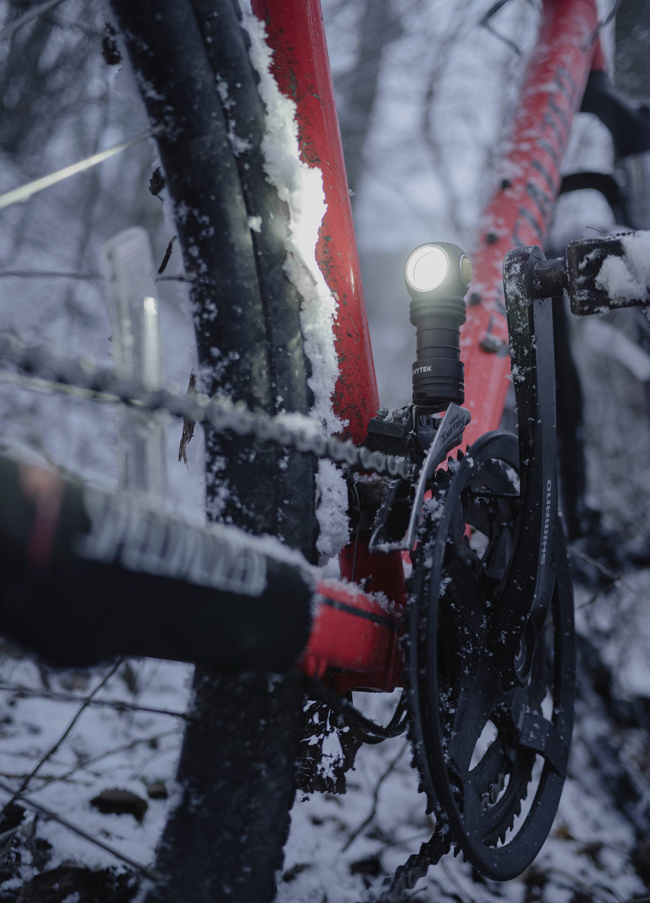 Nahaufnahme eines roten Fahrrads im Schnee. Ein eingeschaltetes Licht ist am Rahmen befestigt. Der Hintergrund zeigt einen bewaldeten Bereich.