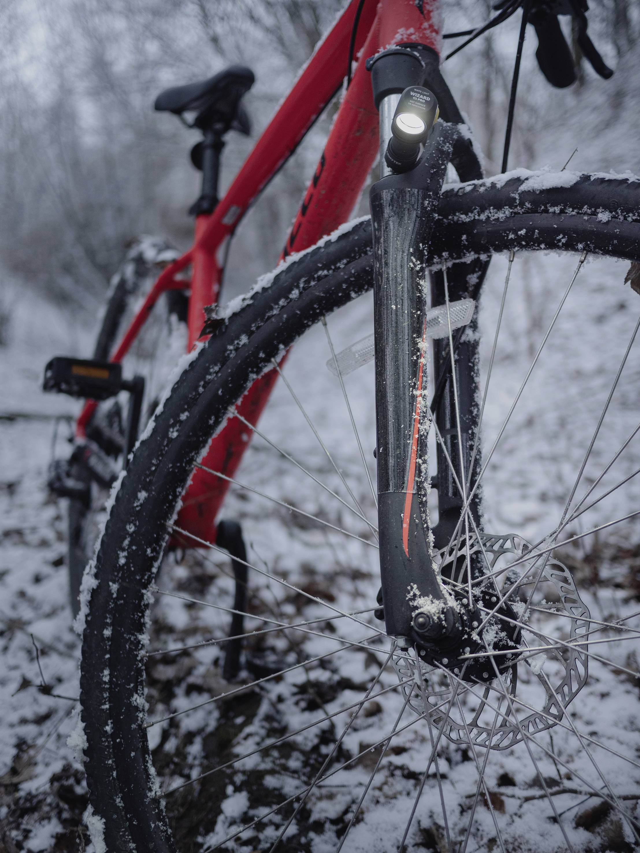 Ein rotes Fahrrad steht im verschneiten Wald. Der Fokus liegt auf dem Vorderreifen und dem montierten Licht. Es ist Winter.