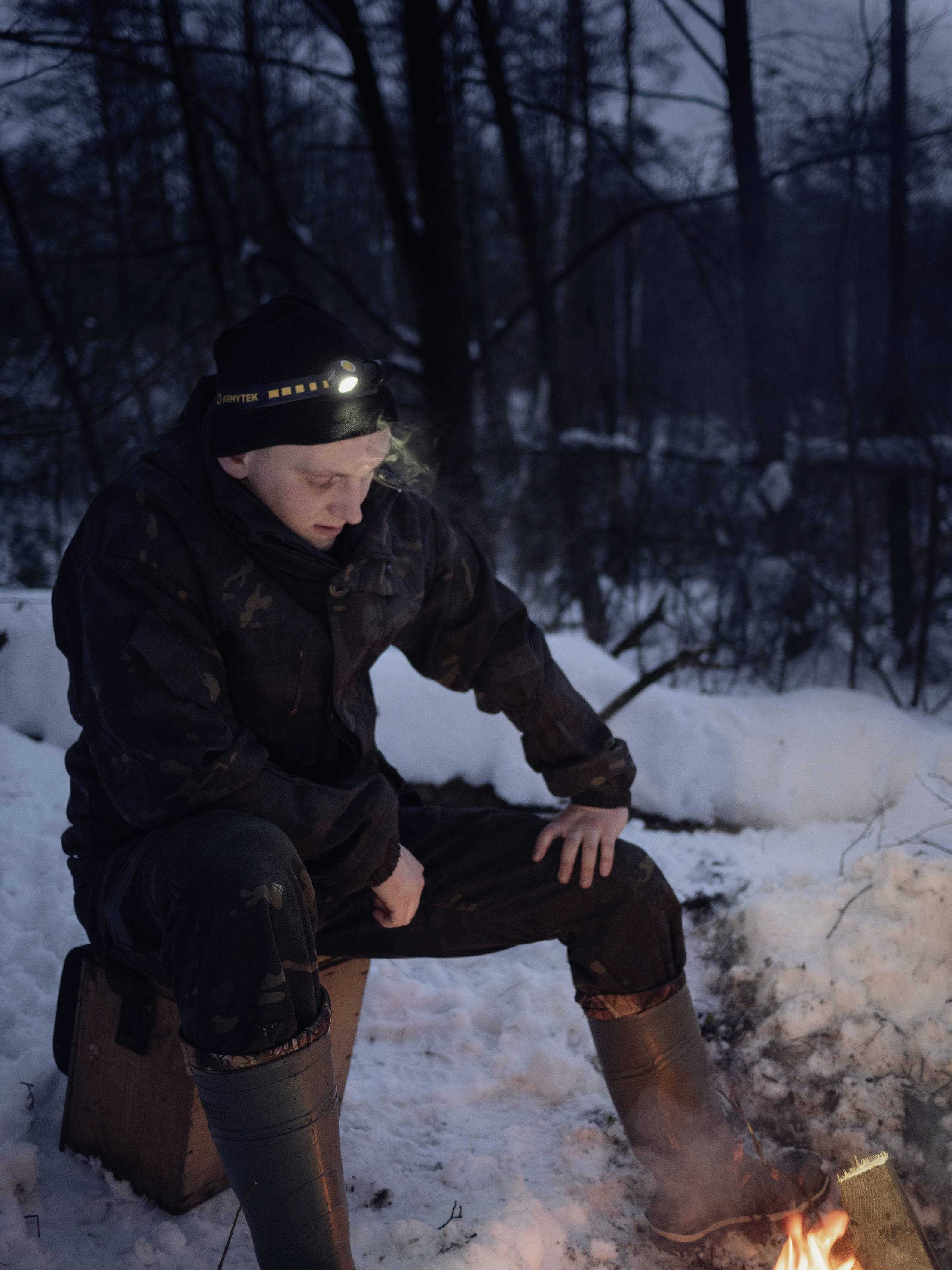 Eine Person sitzt im Schnee mit Winterkleidung neben einem kleinen Lagerfeuer. Abendszene in einem schneebedeckten Wald.