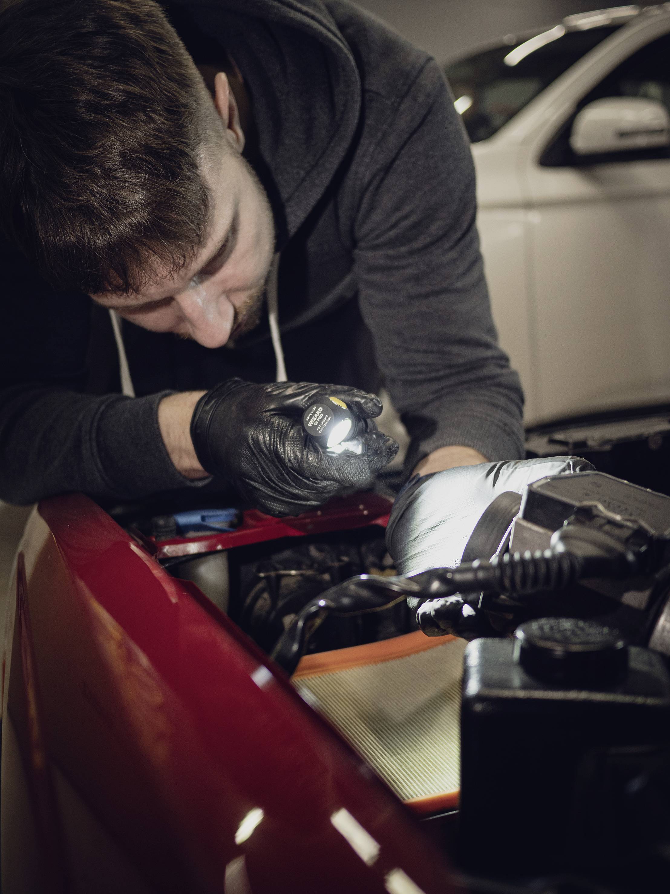 Ein Mechaniker untersucht einen Motor in einer Werkstatt. Er trägt schwarze Handschuhe und verwendet eine Lampe zur Inspektion. Im Hintergrund ist ein weißes Auto zu sehen.