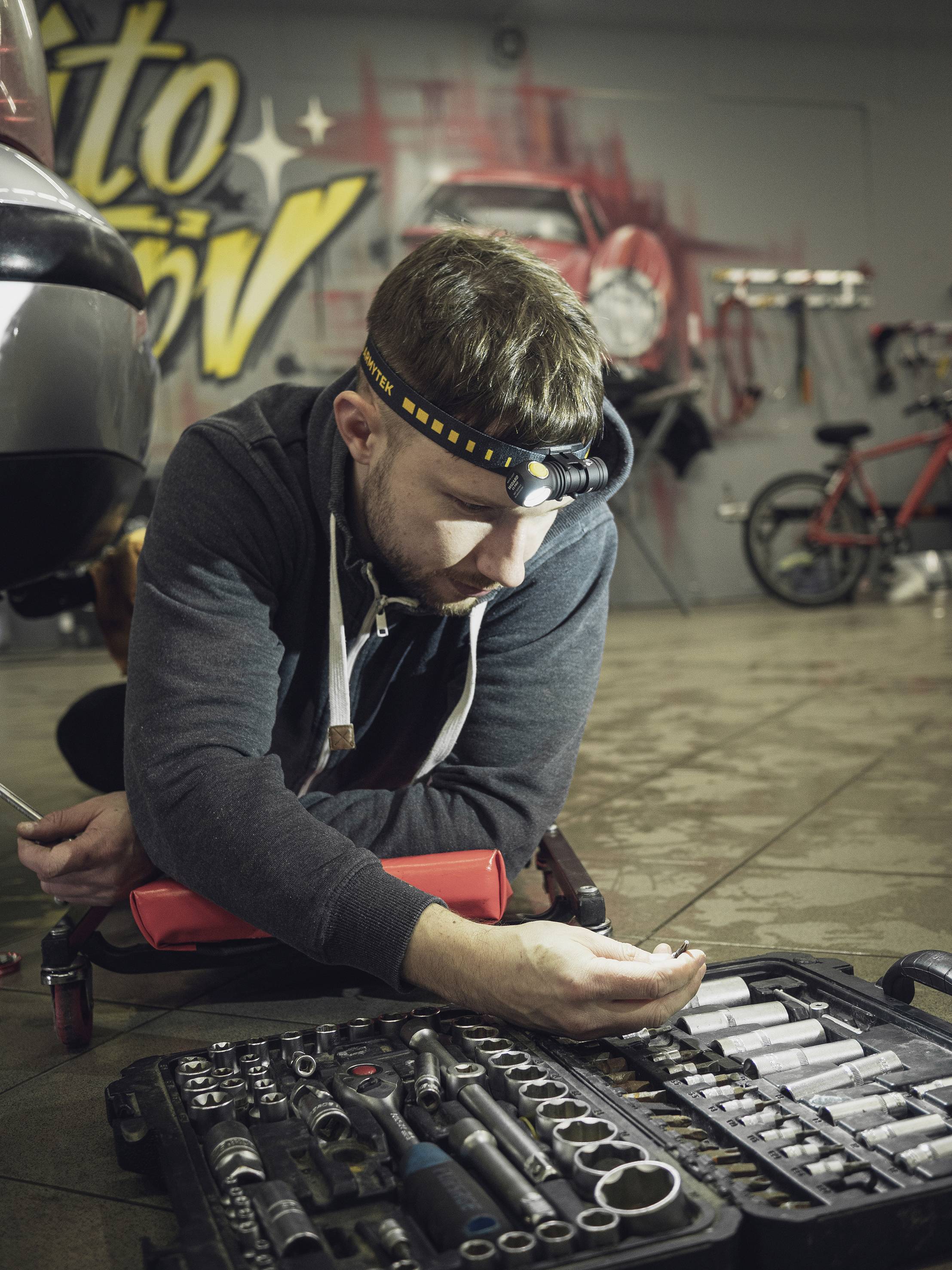 Ein Mechaniker kniet in einer Werkstatt neben einem Werkzeugkasten und überprüft eine kleine Komponente. Im Hintergrund ist ein rotes Fahrrad.
