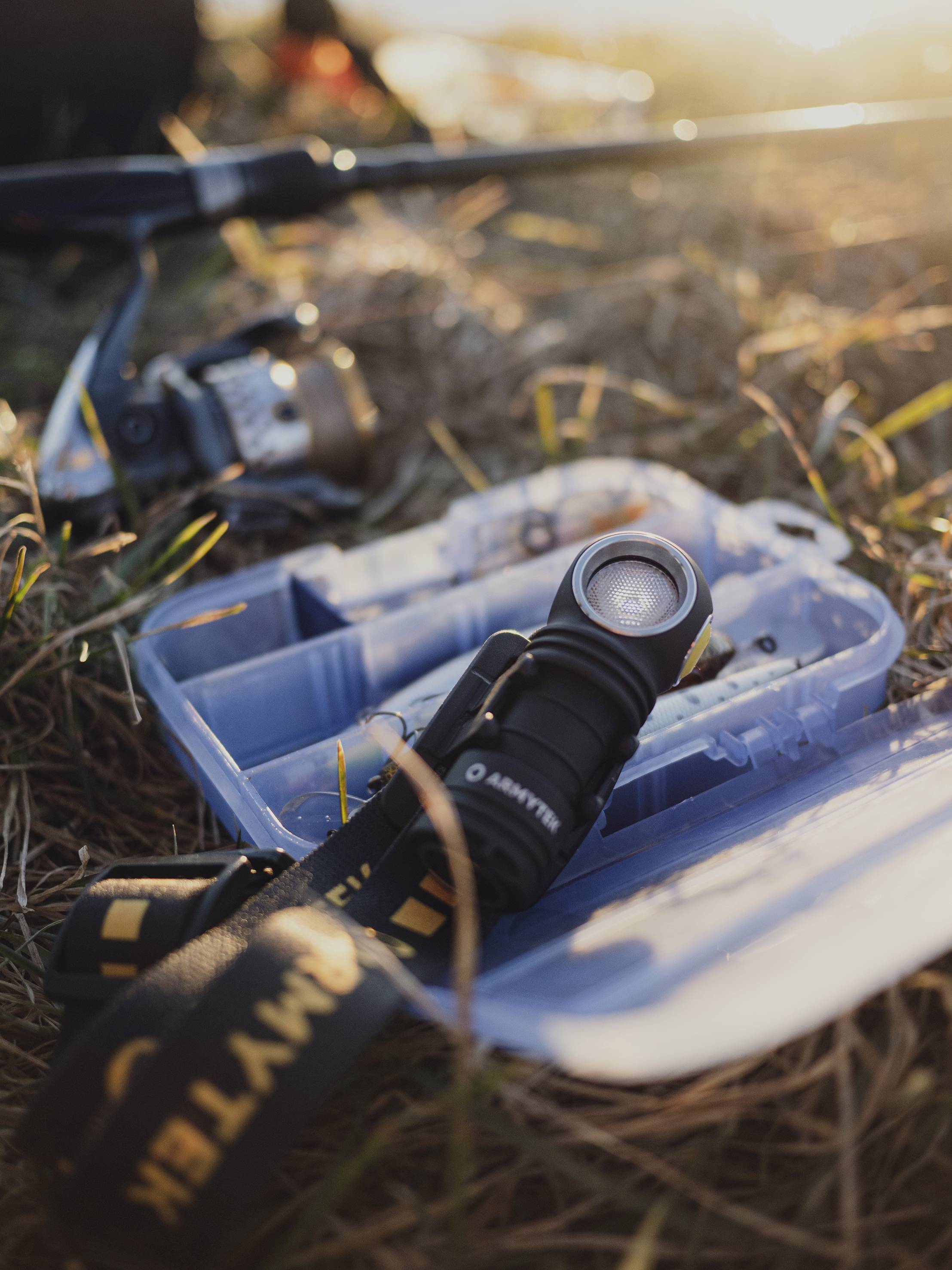 Angeltasche mit Ruten und Spule liegt geöffnet auf Gras im Sonnenuntergang; Taschenlampe im Vordergrund.