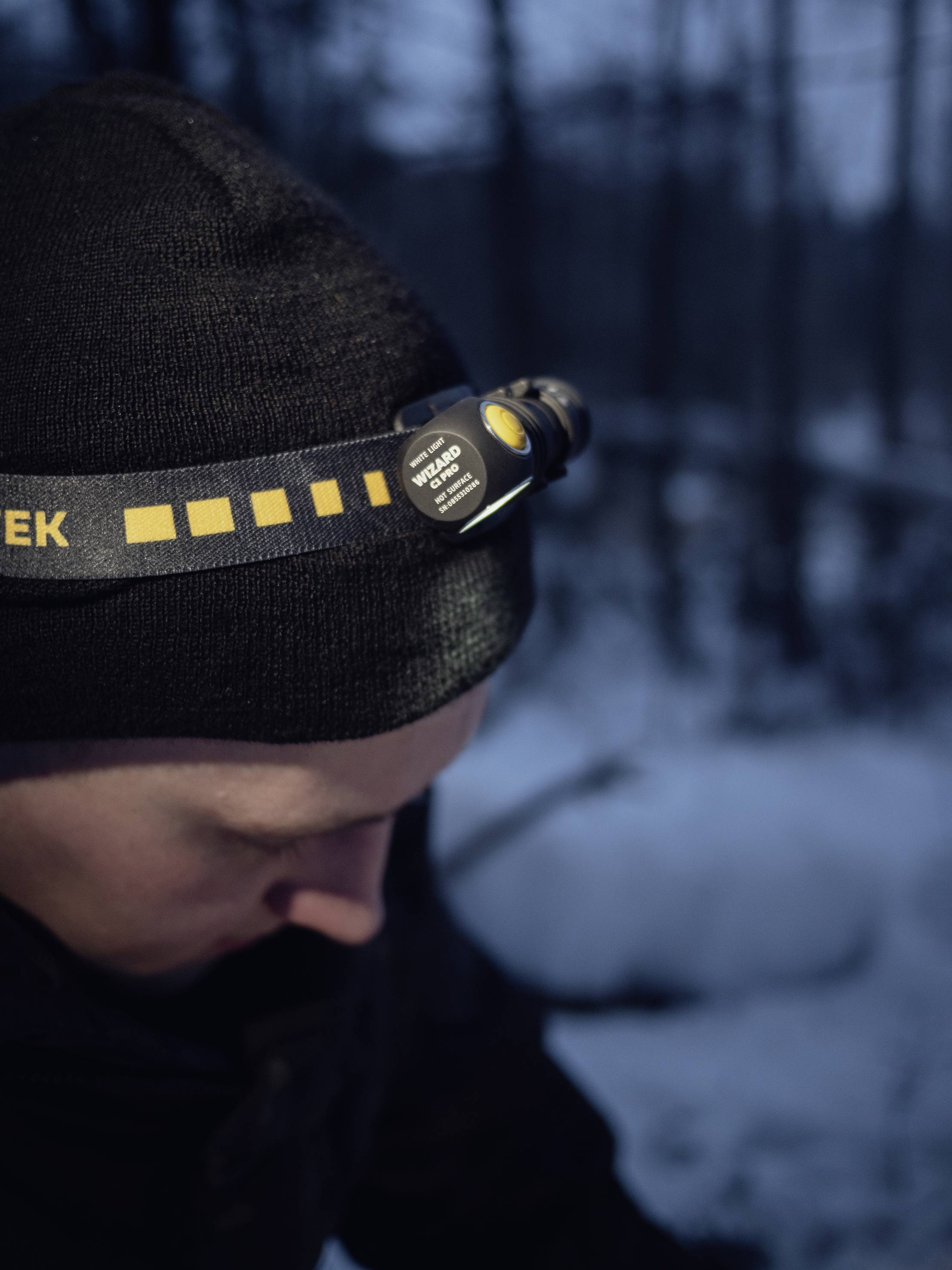 Eine Person trägt eine schwarze Mütze mit einer Stirnlampe, draußen in einer schneebedeckten Waldlandschaft bei Dämmerung.